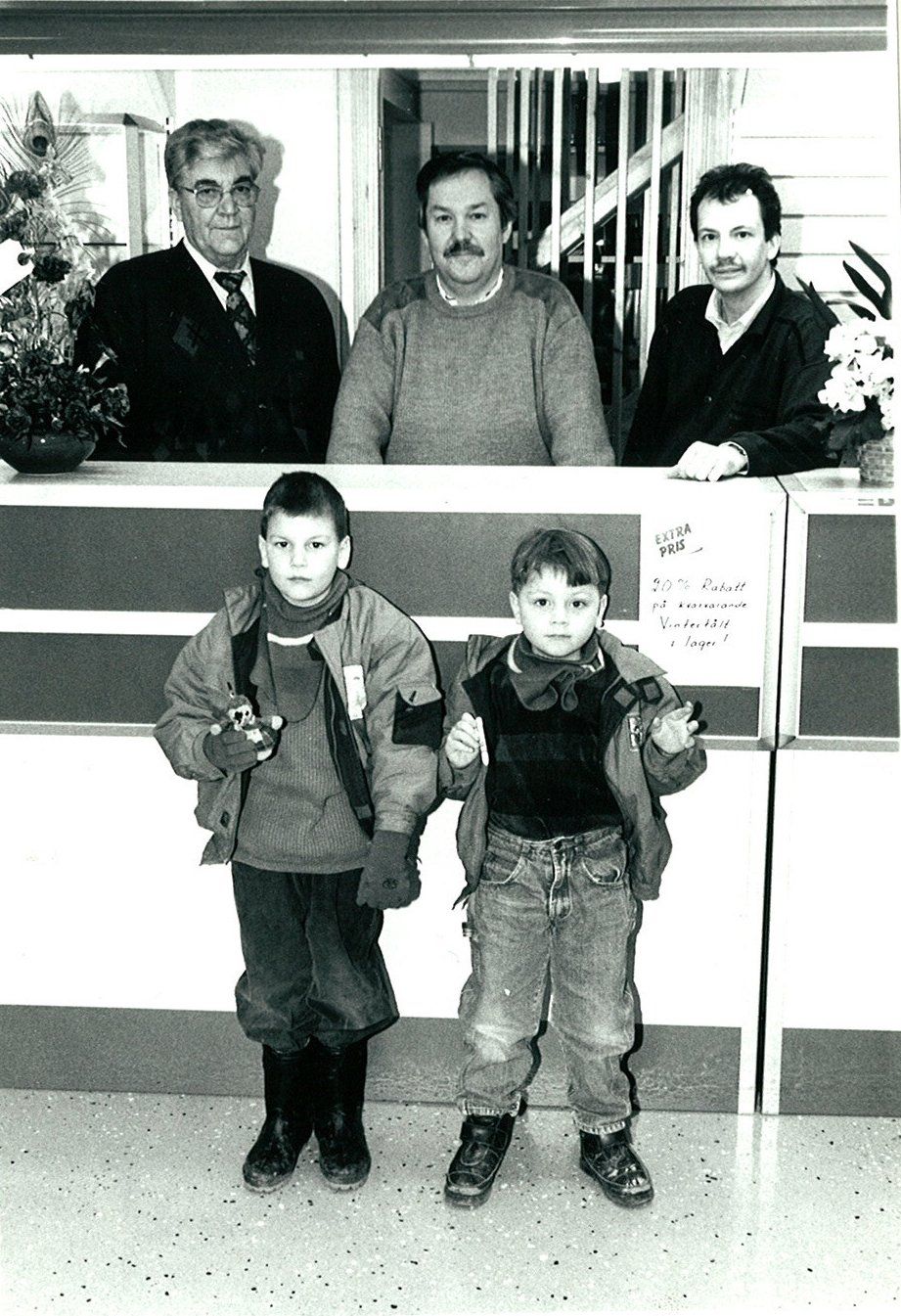 Two young boys pose in front of a counter with three men standing behind it.