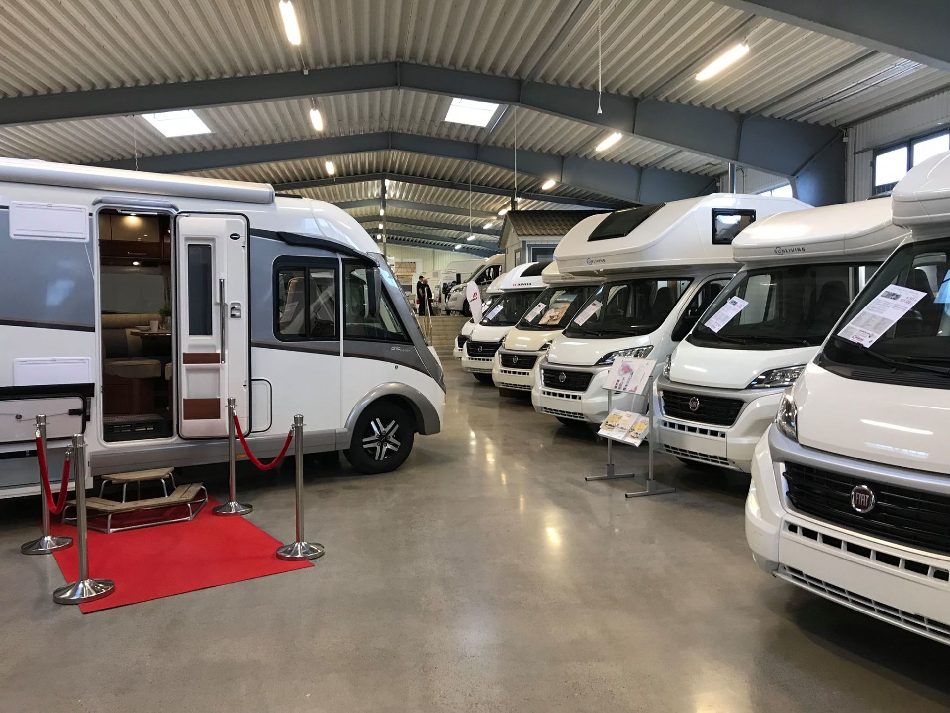 Inside RV showroom: Several white and gray recreational vehicles displayed on a concrete floor, red carpet in front.