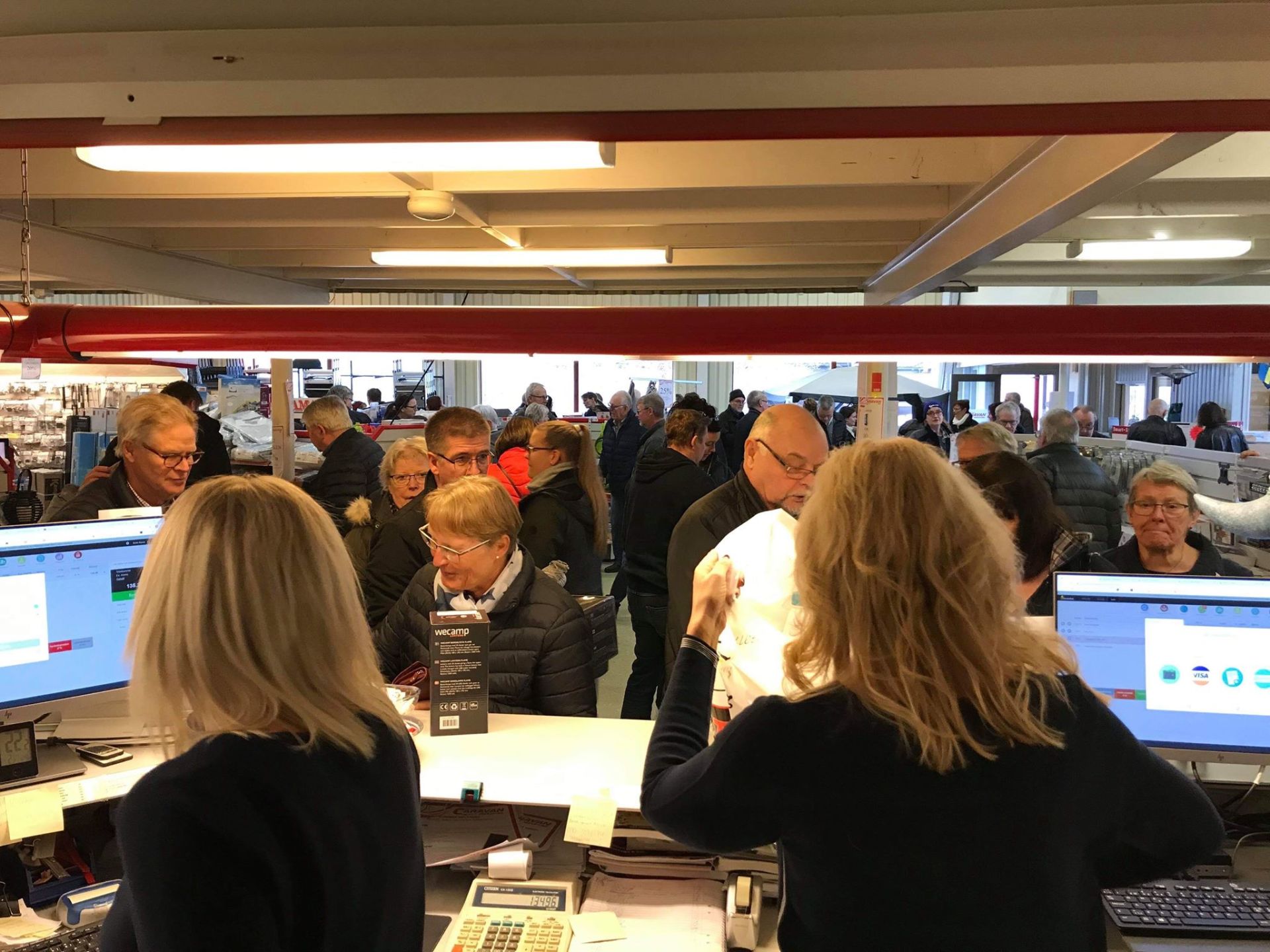 People at a busy counter; two women behind it, customers in a queue, possibly at a store.