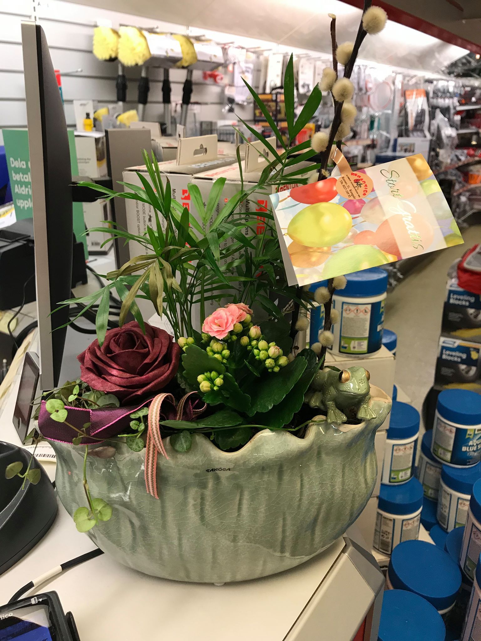 Floral arrangement in a green, textured ceramic pot, with a greeting card in a retail setting.