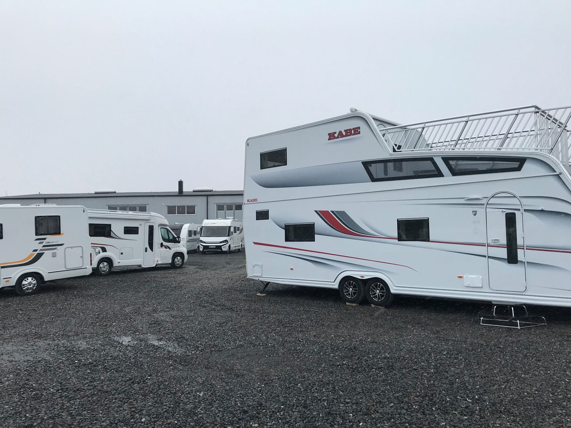 Campers parked on gravel lot. White and colored RVs, overcast sky. Industrial building in the background.