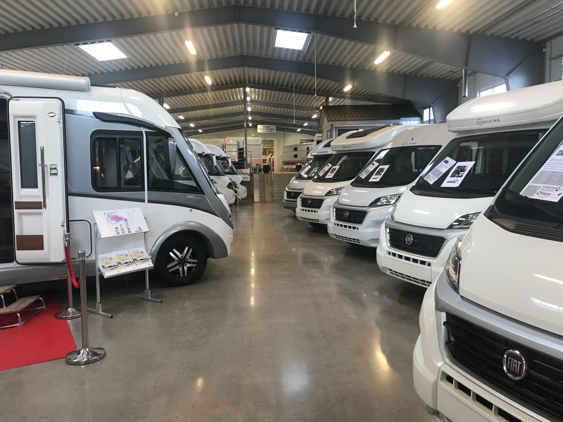 RVs parked inside a large, bright showroom. Several white and grey campers displayed in rows.
