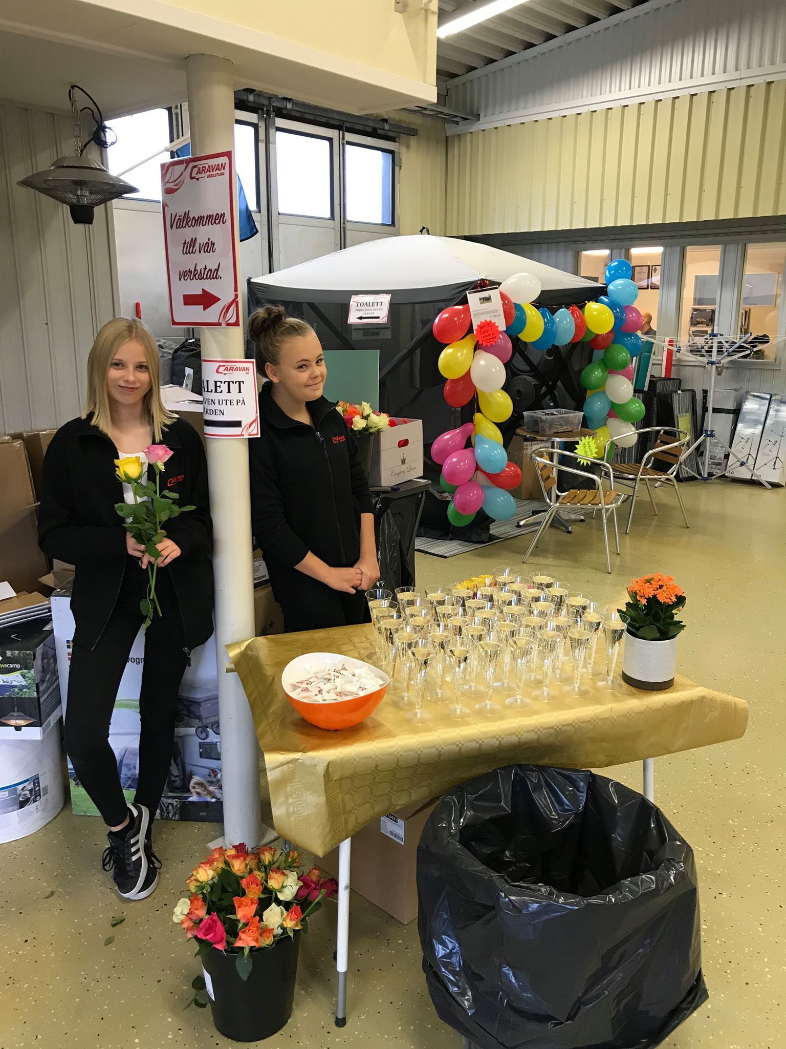 Two young women at a party, one holding a rose. A table with food and drinks, balloons, and decorations are present.