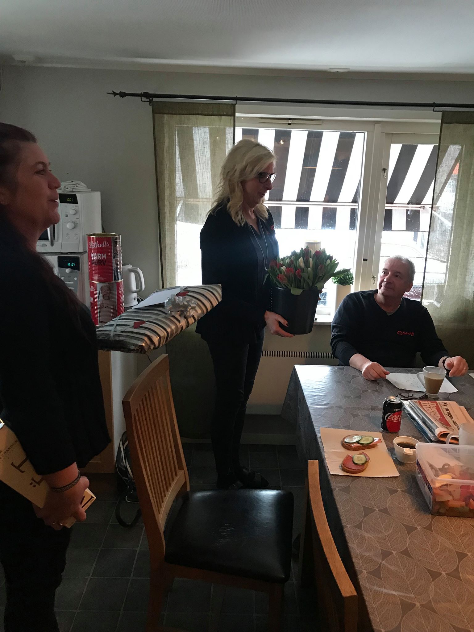 Woman holding flowers, standing near a table with a man and another woman in a room with a window.