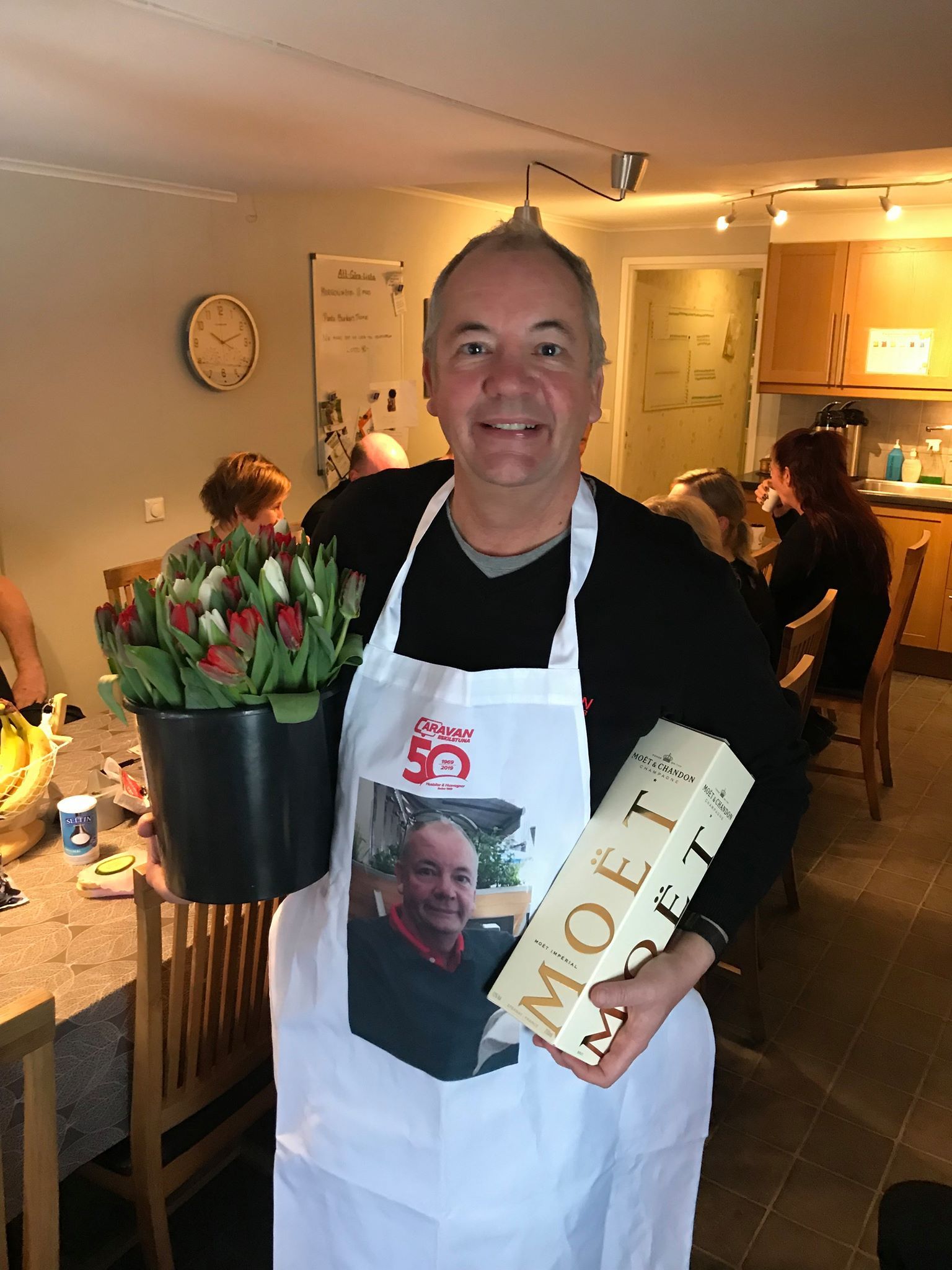 Man in apron holding flowers and champagne, smiling in a room.