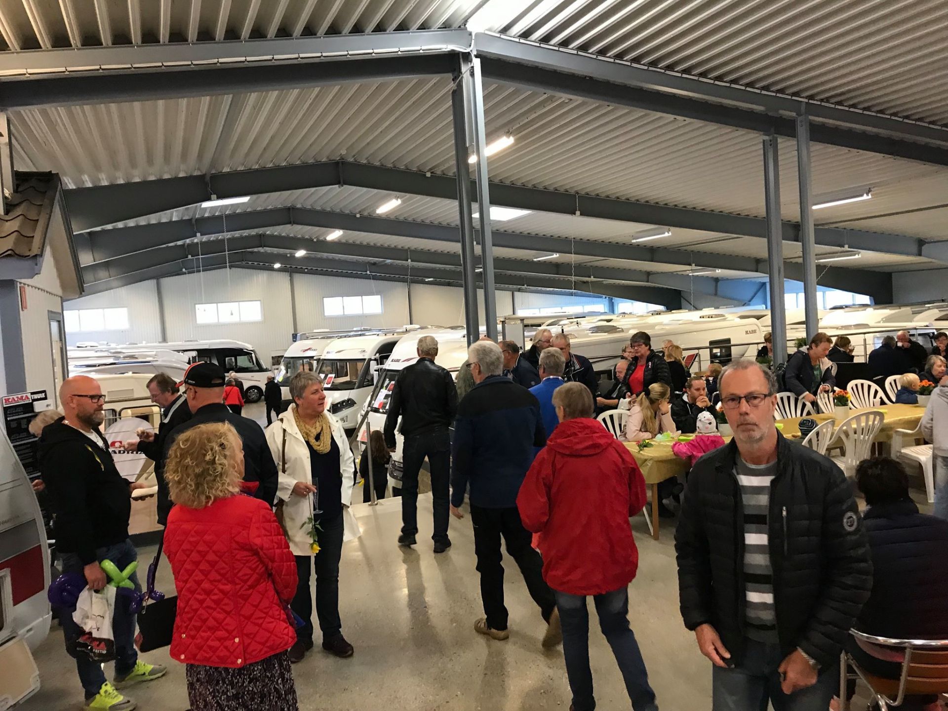 People gathered indoors; RVs visible in the background. Many are socializing, some at tables.