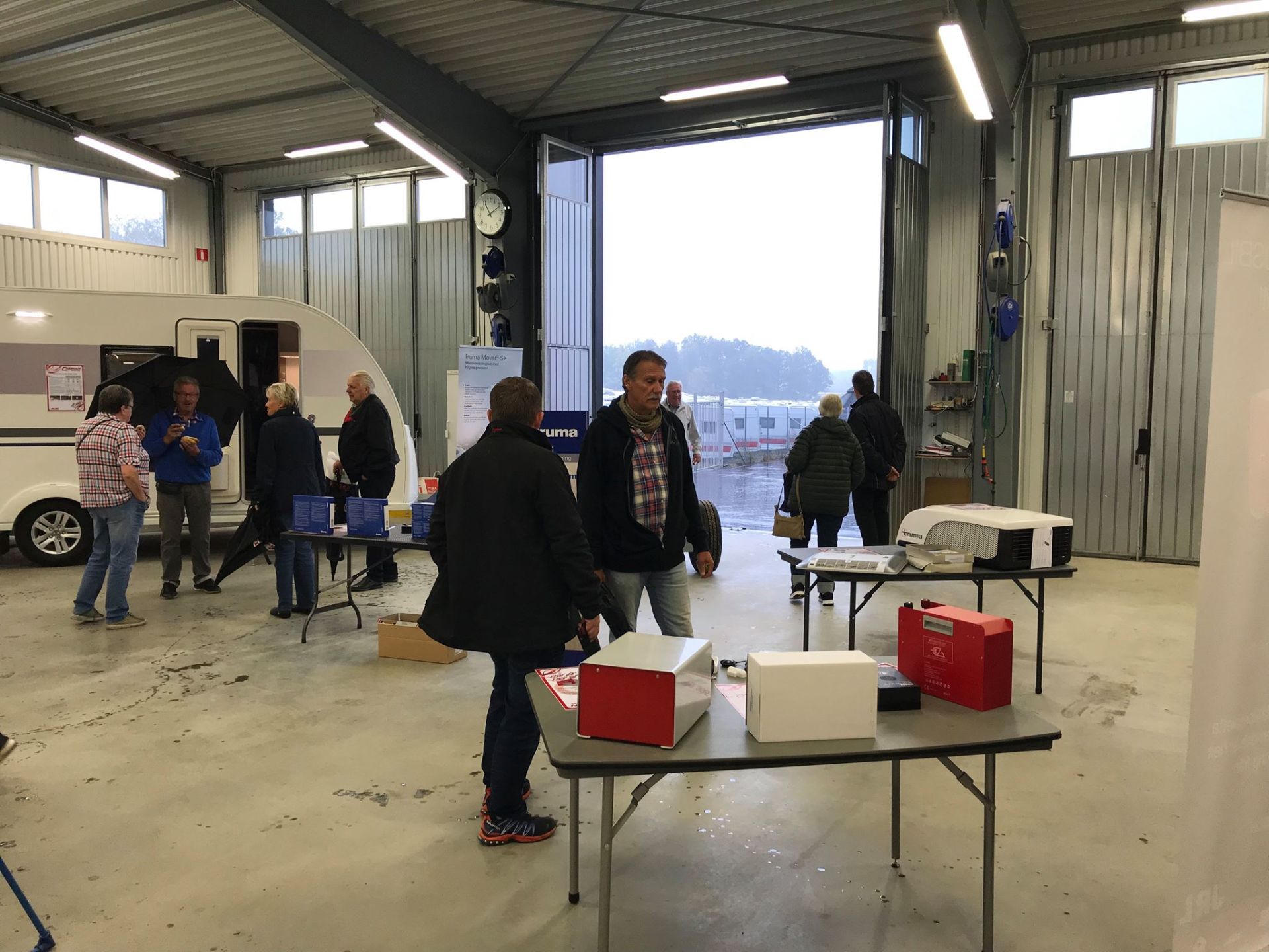 People gathered inside a garage, near tables with boxes and a camper. The door is open to an exterior view.