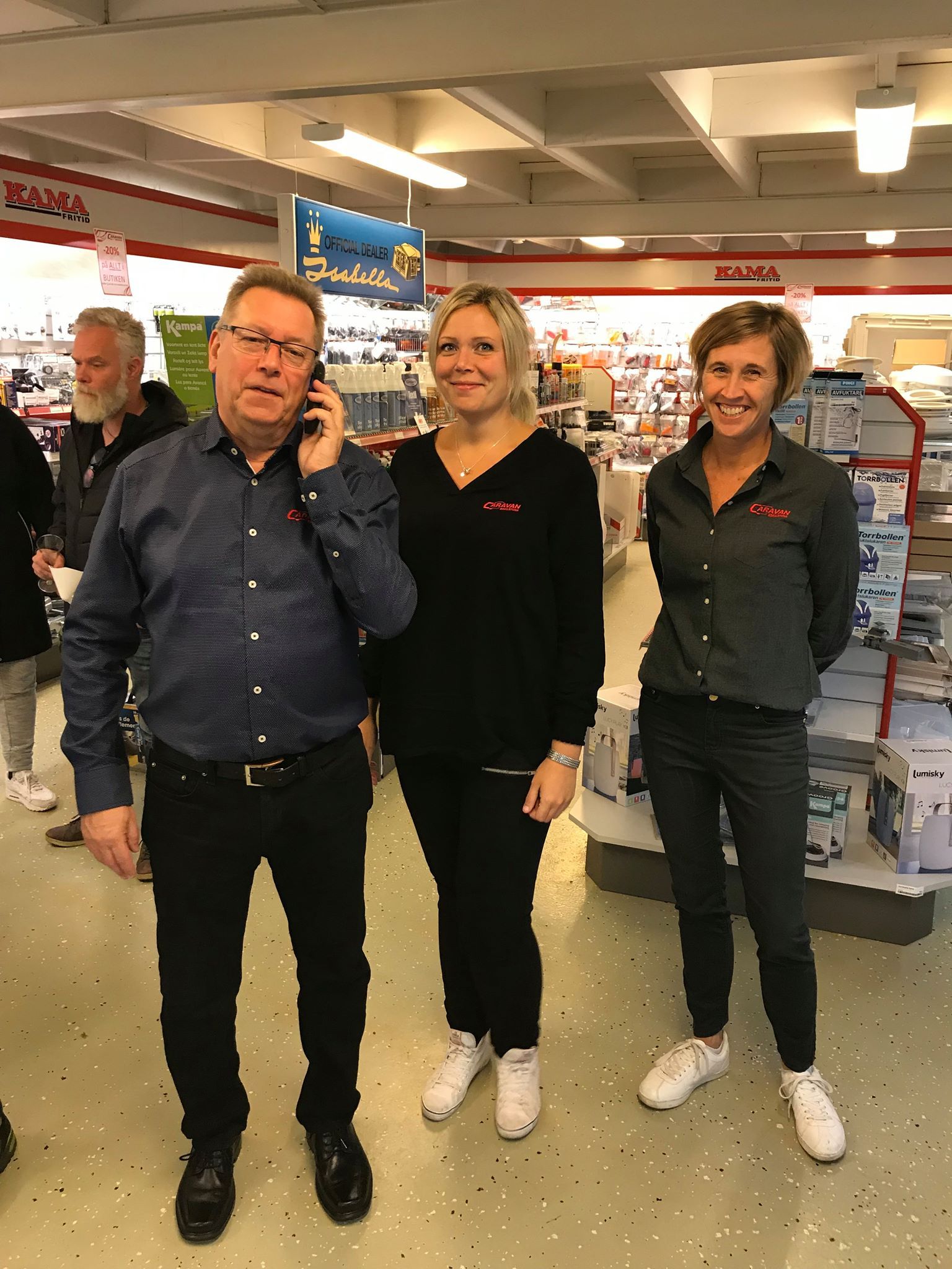 A man on a phone, flanked by two women, in a store. All three wear company branded clothing.