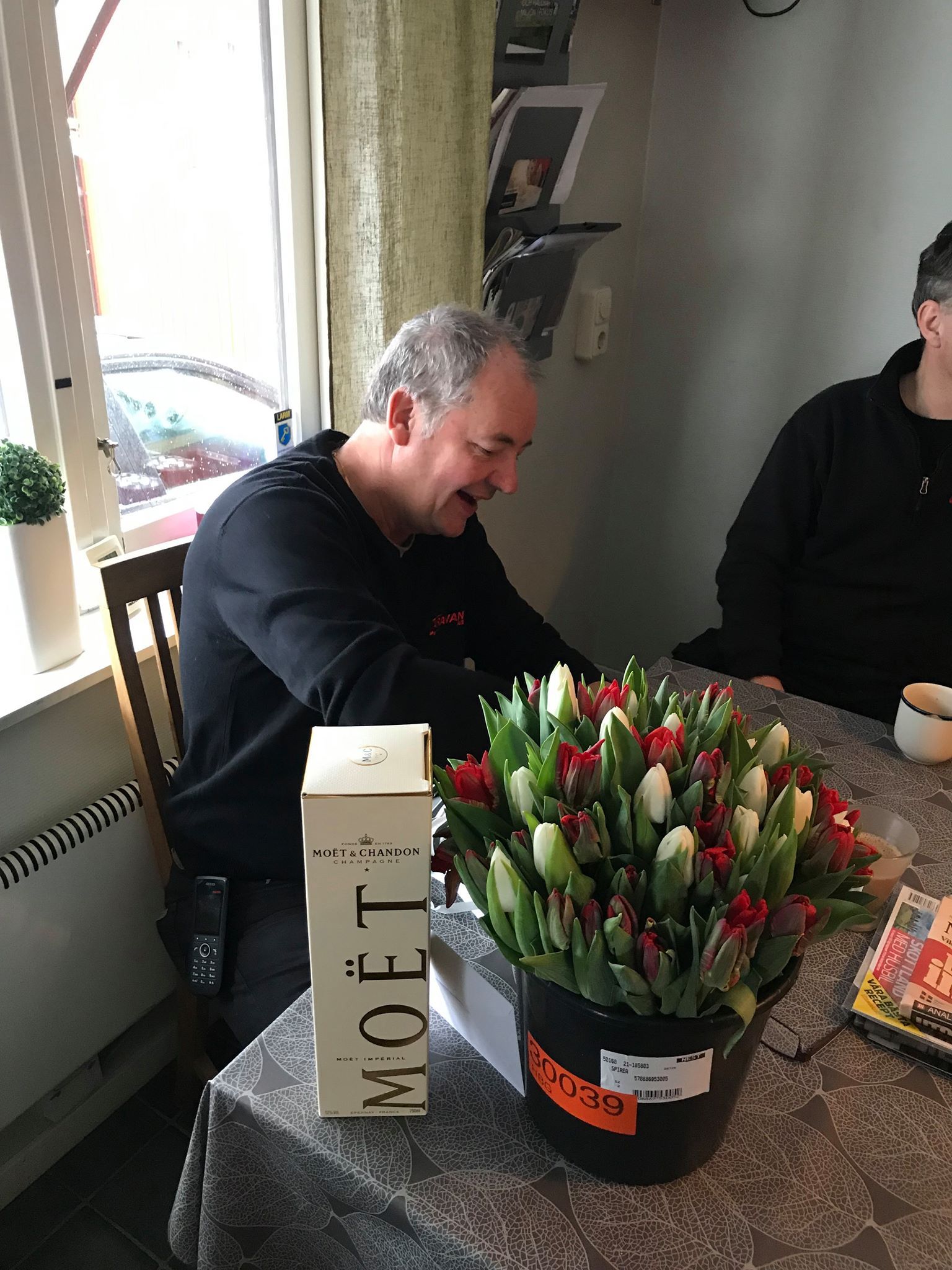 Man at table with flowers and Moët box. He's smiling. Interior shot with window.