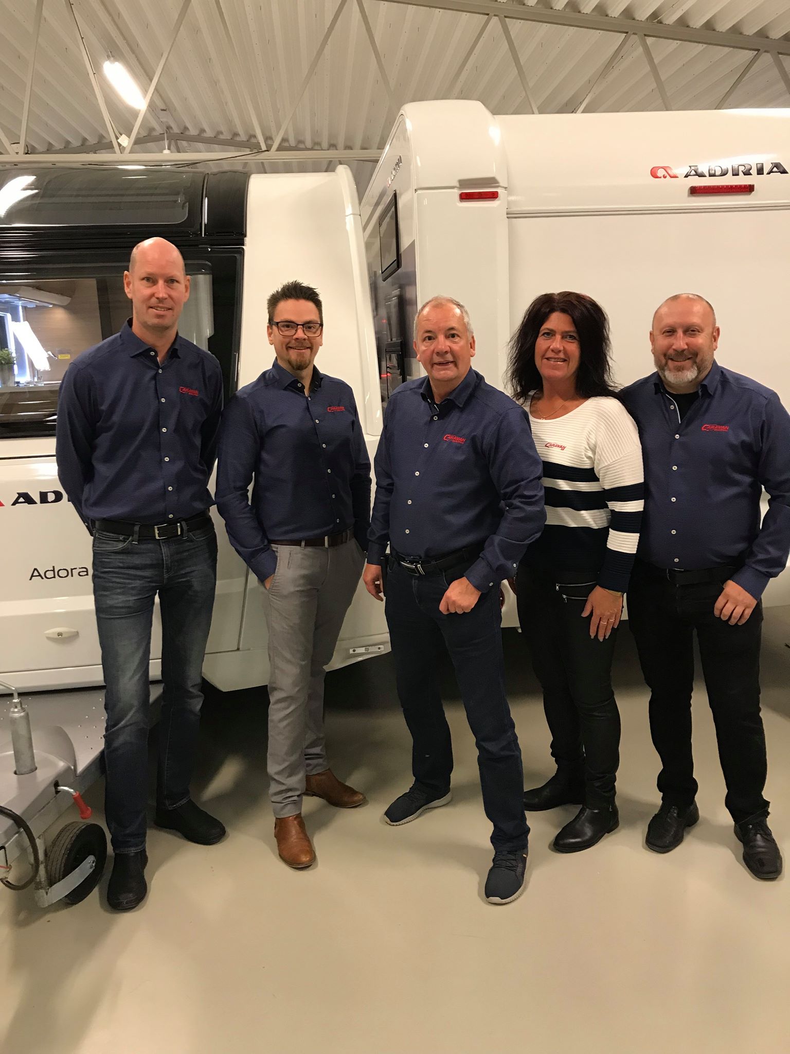 Five people standing in front of a white camper. They wear dark blue shirts and black pants, smiling in a well-lit showroom.