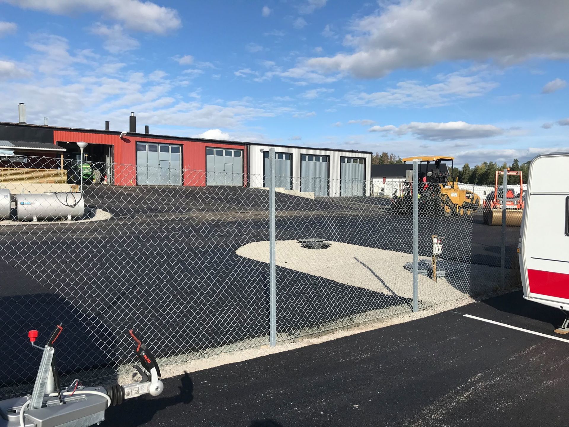Black asphalt parking lot, chain-link fence, industrial building with red and silver garage doors, clear sky.