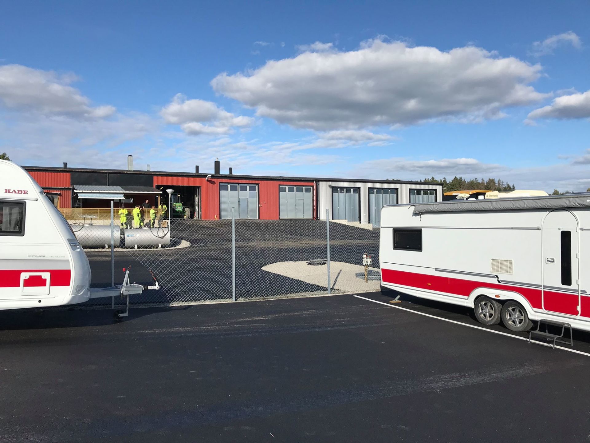 Caravans parked near a red and gray storage building under a blue sky with clouds.