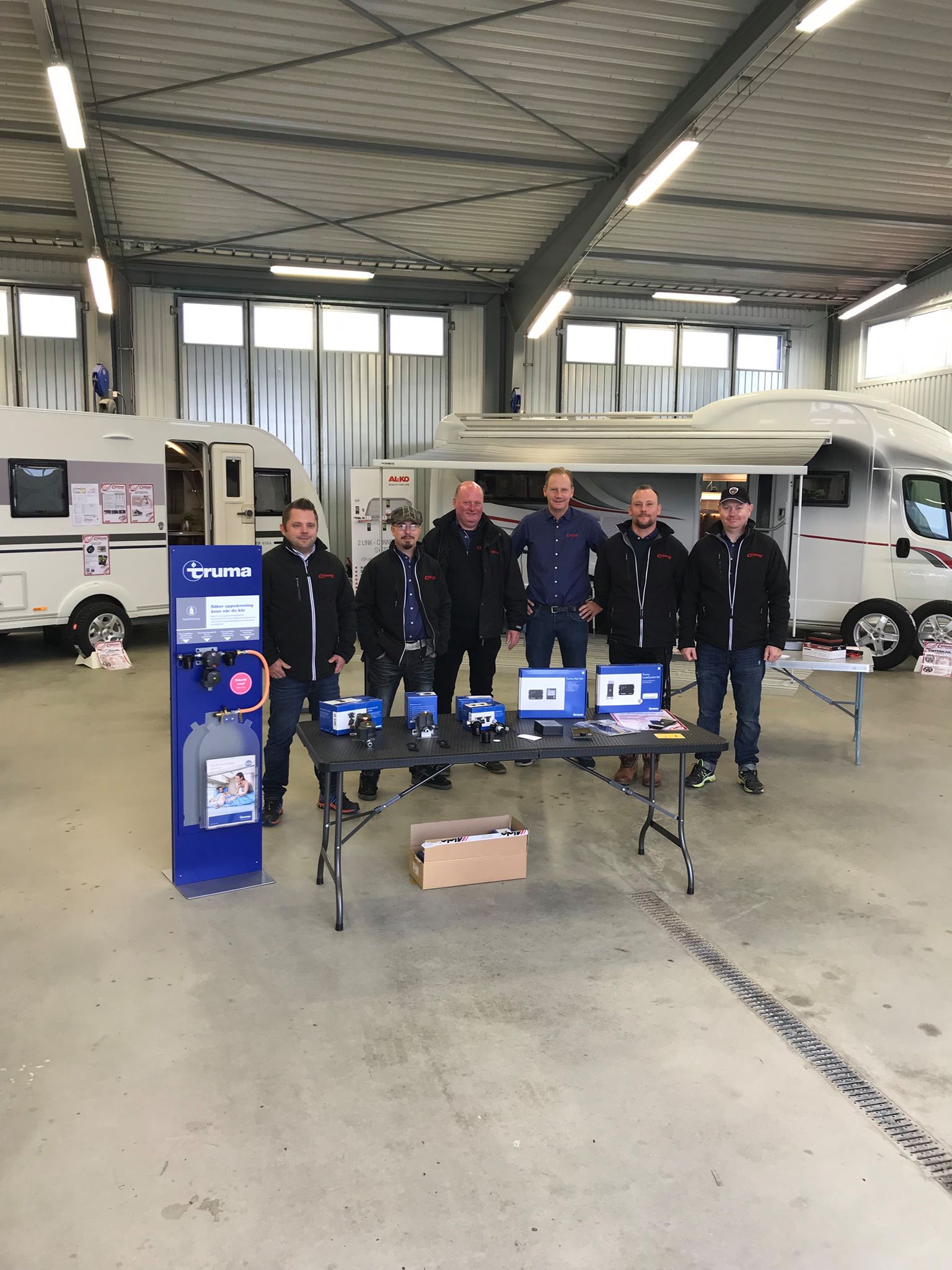 Group of men in a garage, standing behind tables with electronic devices, RVs in background.