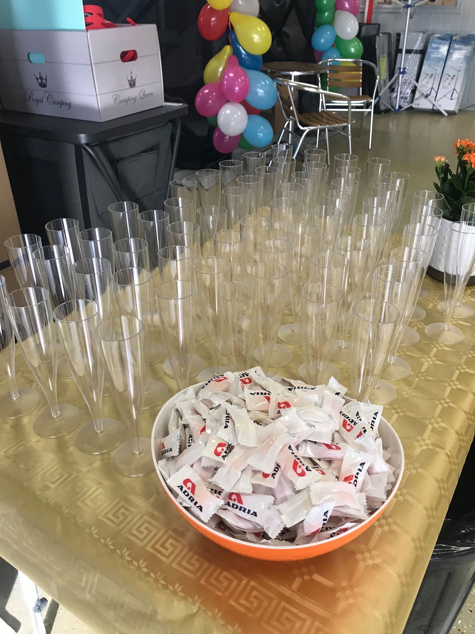 Table set for an event with champagne flutes and bowl of wrapped candies; balloons and boxes in background.