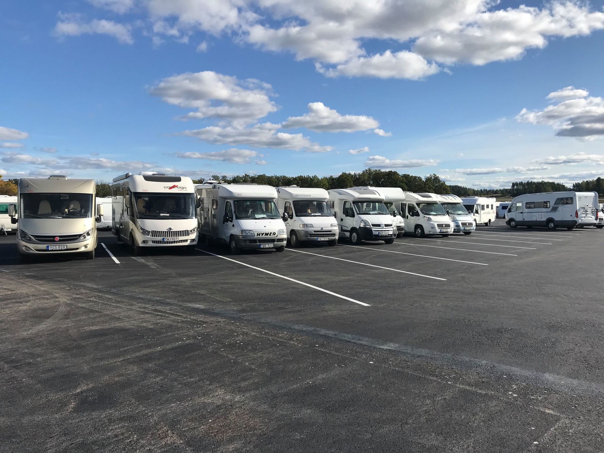 Several white RVs parked in an asphalt lot under a blue sky with clouds.