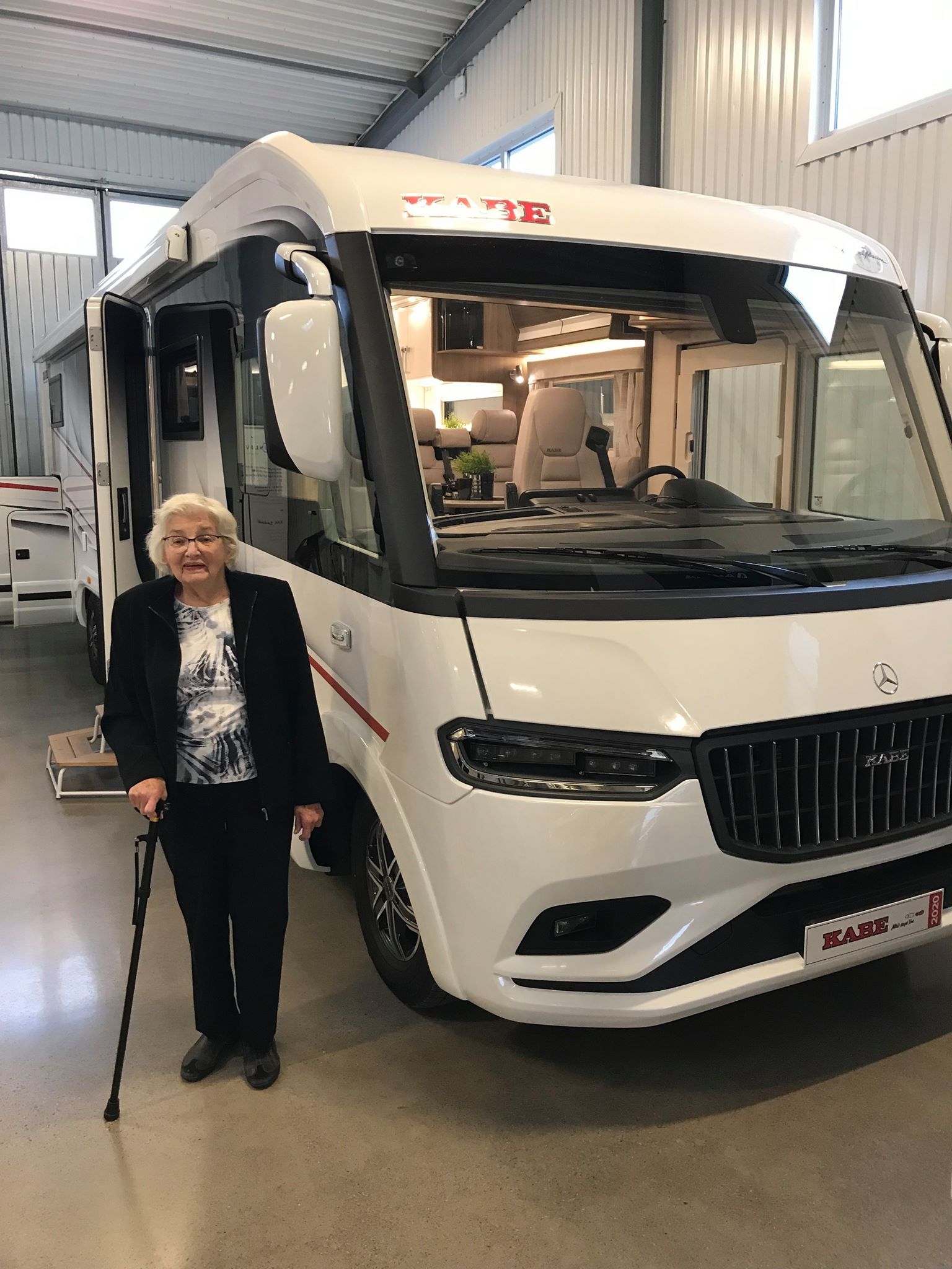 Woman with cane stands in front of a white RV. She smiles, indoors.