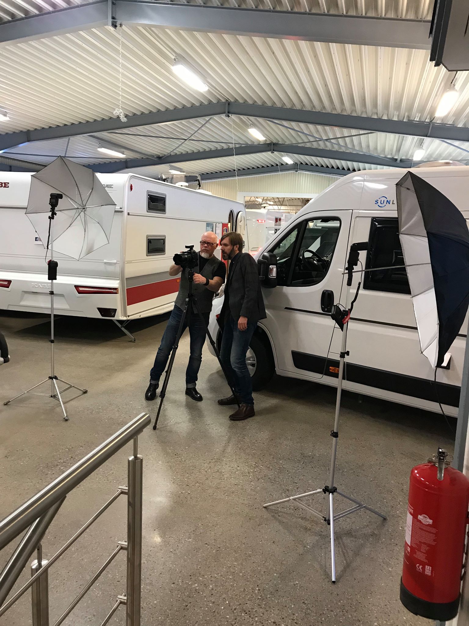 Two men pose beside a white van in a showroom, lit by umbrellas on tripods.