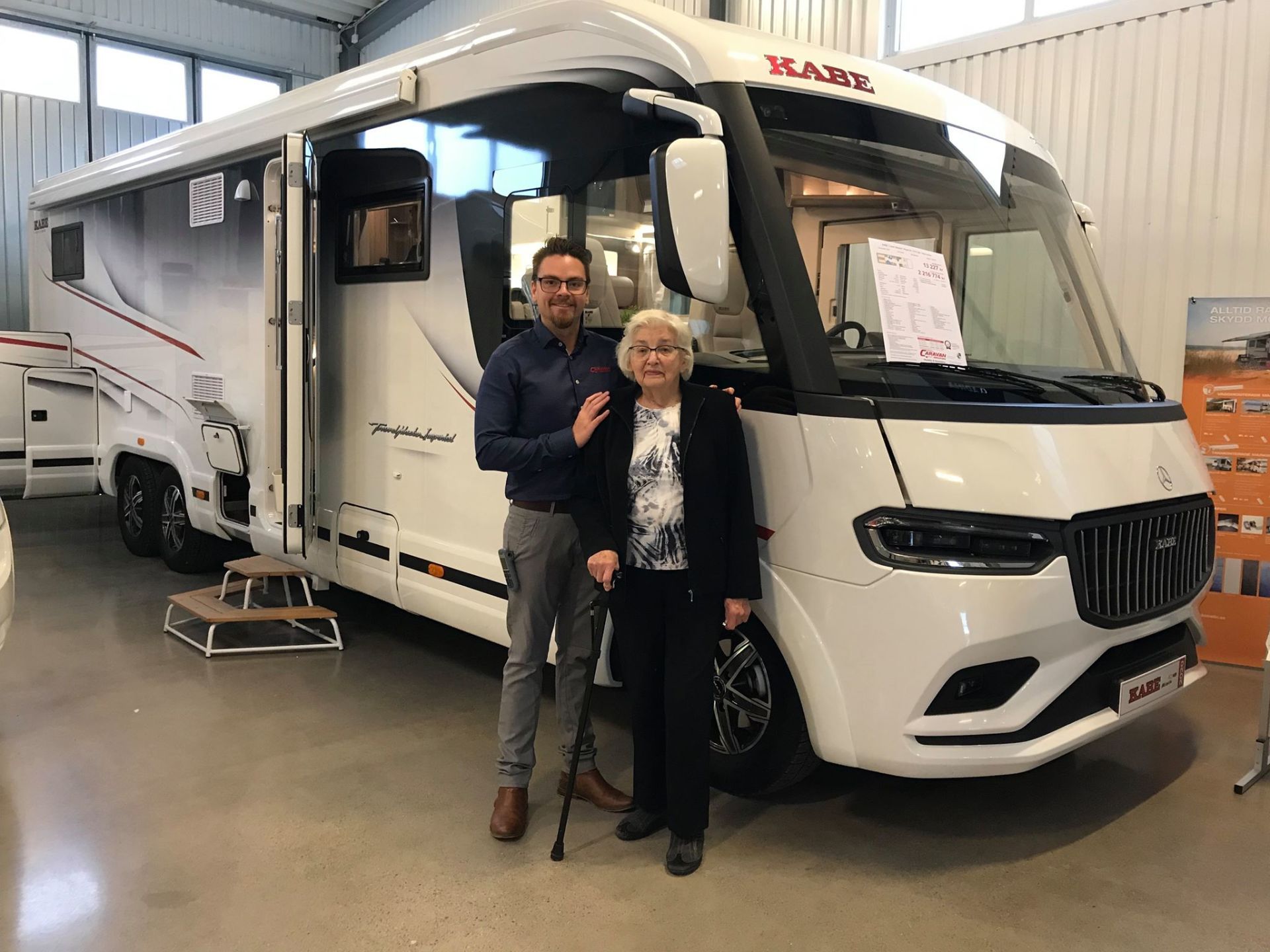 Man and woman posing in front of a white RV. The woman uses a cane. Indoors, showroom.