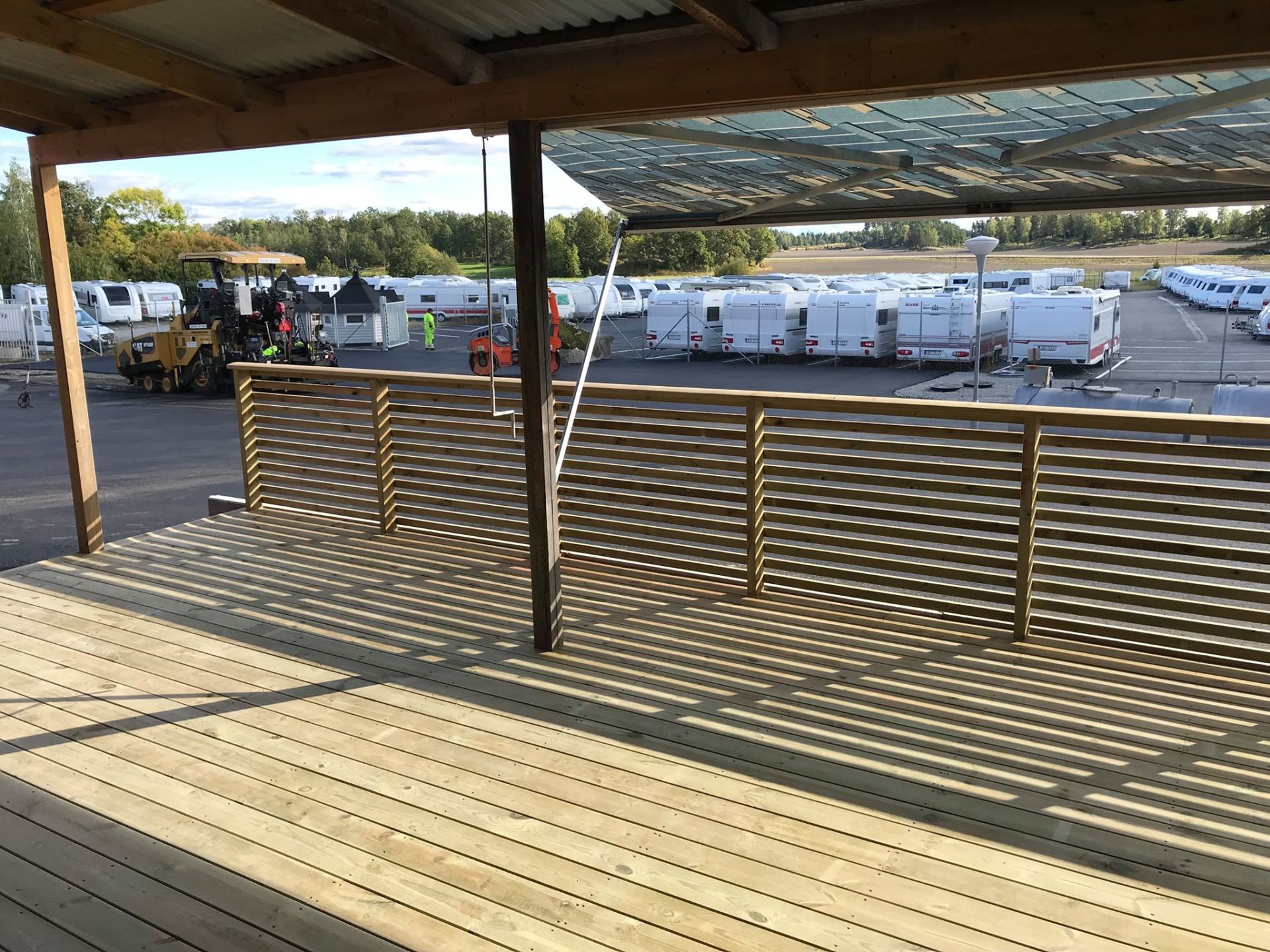 Wooden deck with slatted railing under a corrugated roof, overlooking a parking lot with trailers and construction equipment.