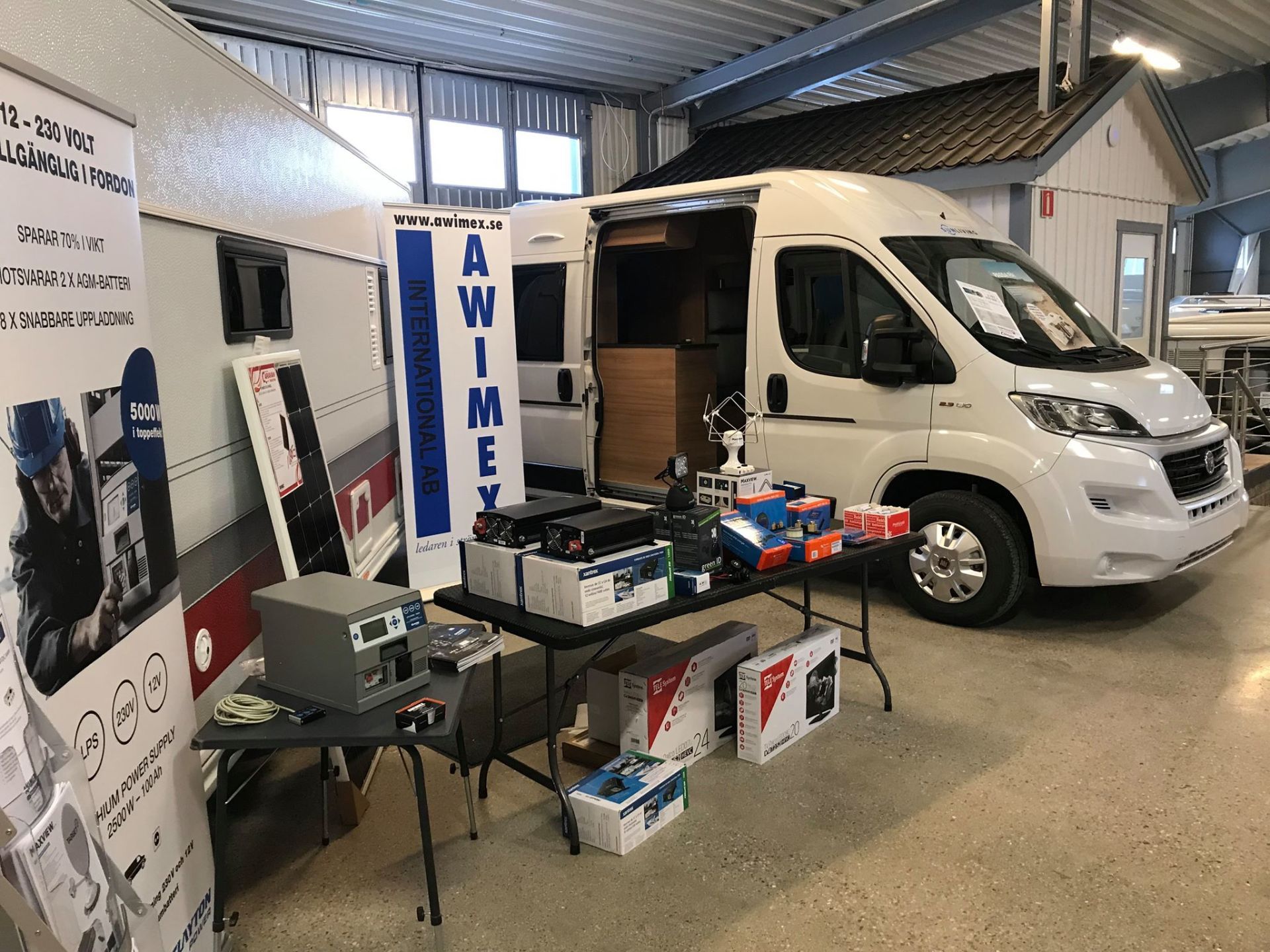 A white camper van on display with related equipment on a table, in a showroom setting.