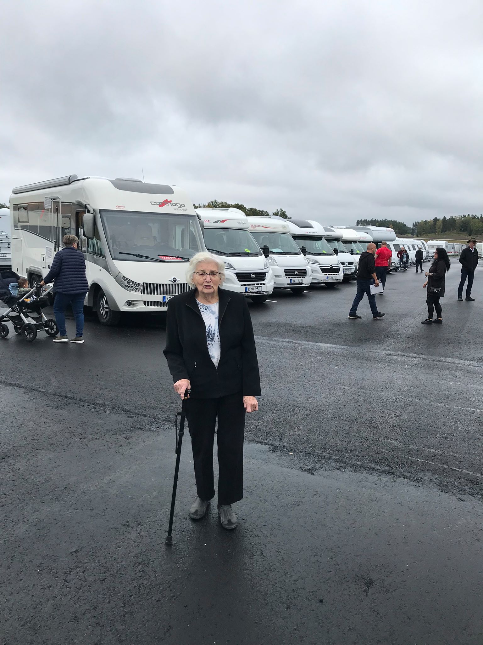 Woman with cane in front of RVs, parked on wet asphalt on an overcast day, people in the background.
