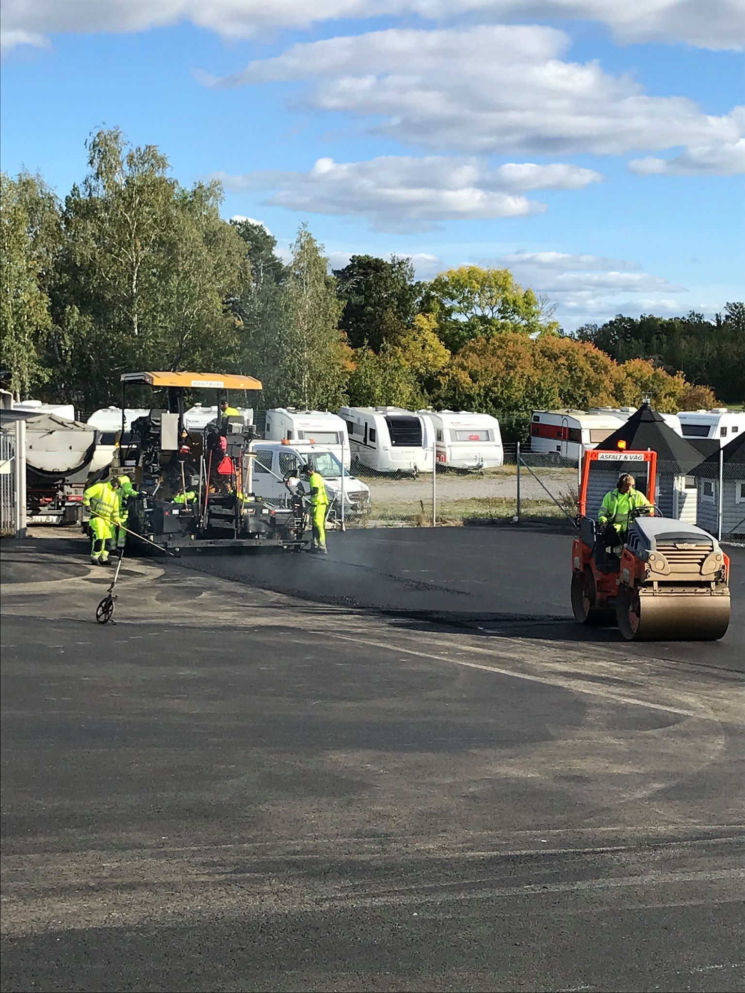 Asphalt paving on a lot. Construction workers in safety vests operate machinery: paver, roller. Trailers parked in background.