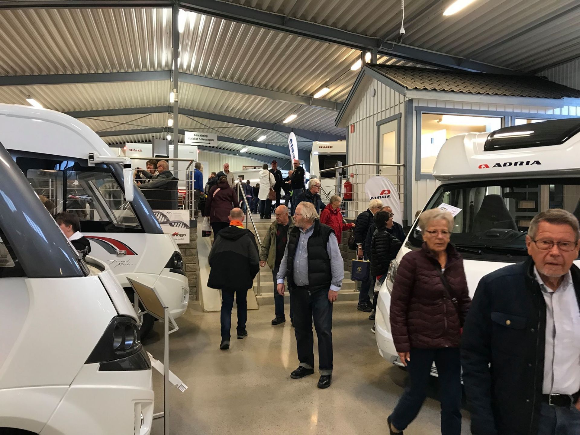 People browse motorhomes inside a large building. White vehicles, a corrugated metal roof, and various vendors are visible.