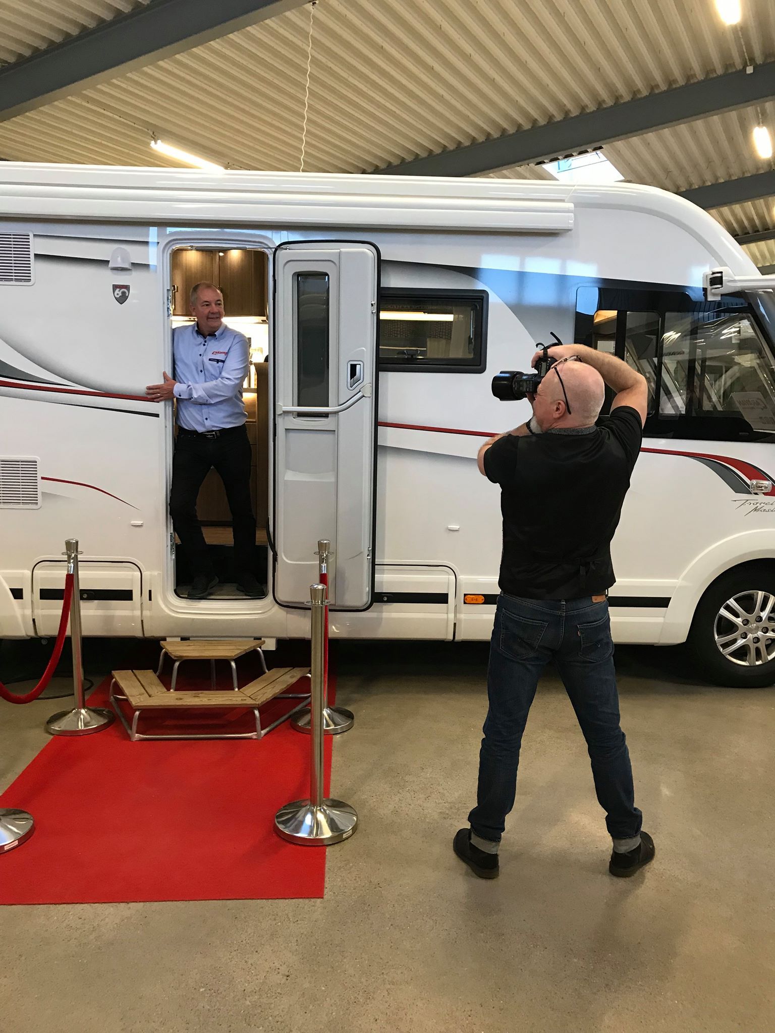 Man poses in RV doorway while photographer takes his picture; red carpet in front.