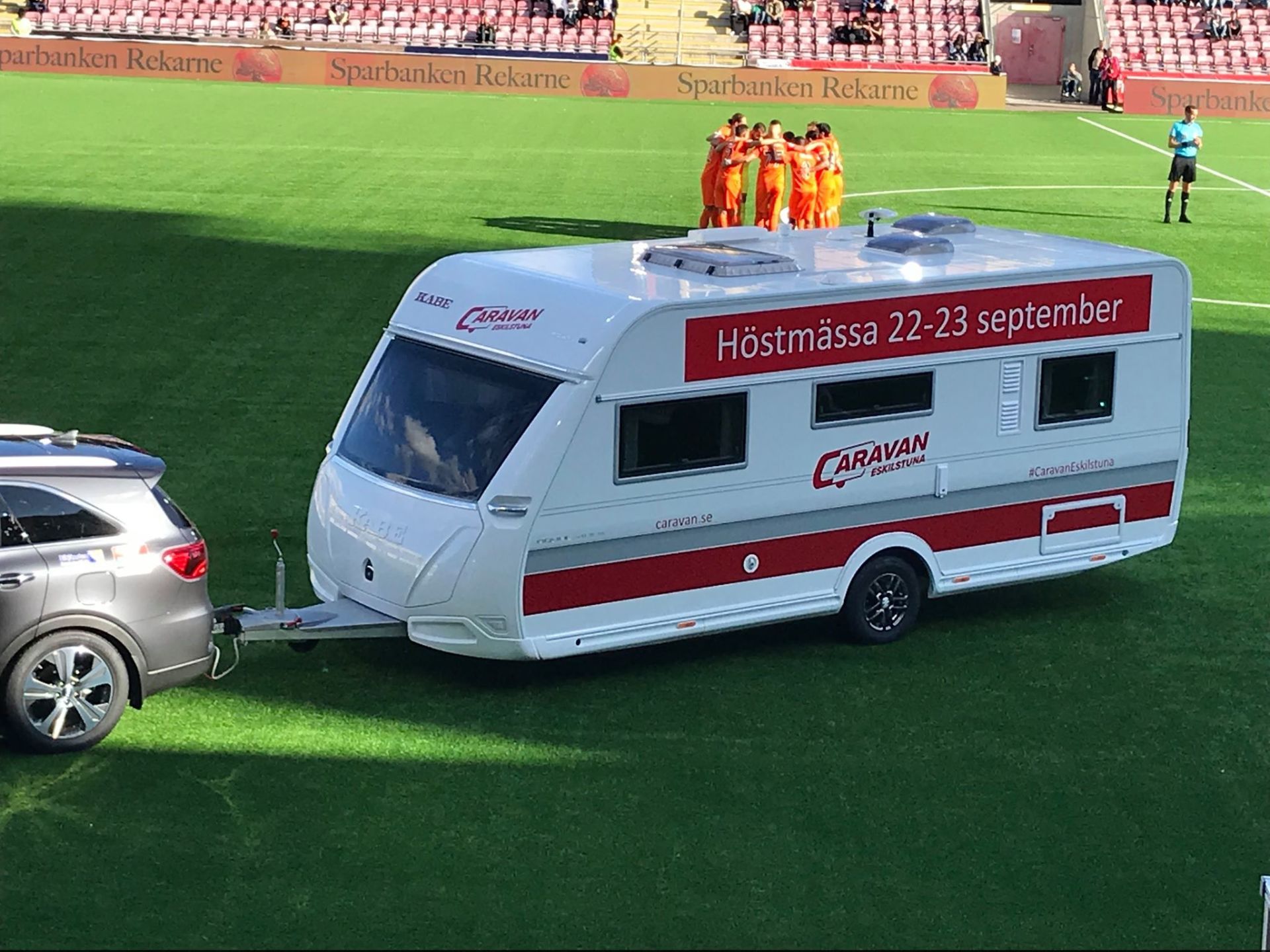 A white Caravan trailer being towed by a gray SUV on a soccer field during a game.