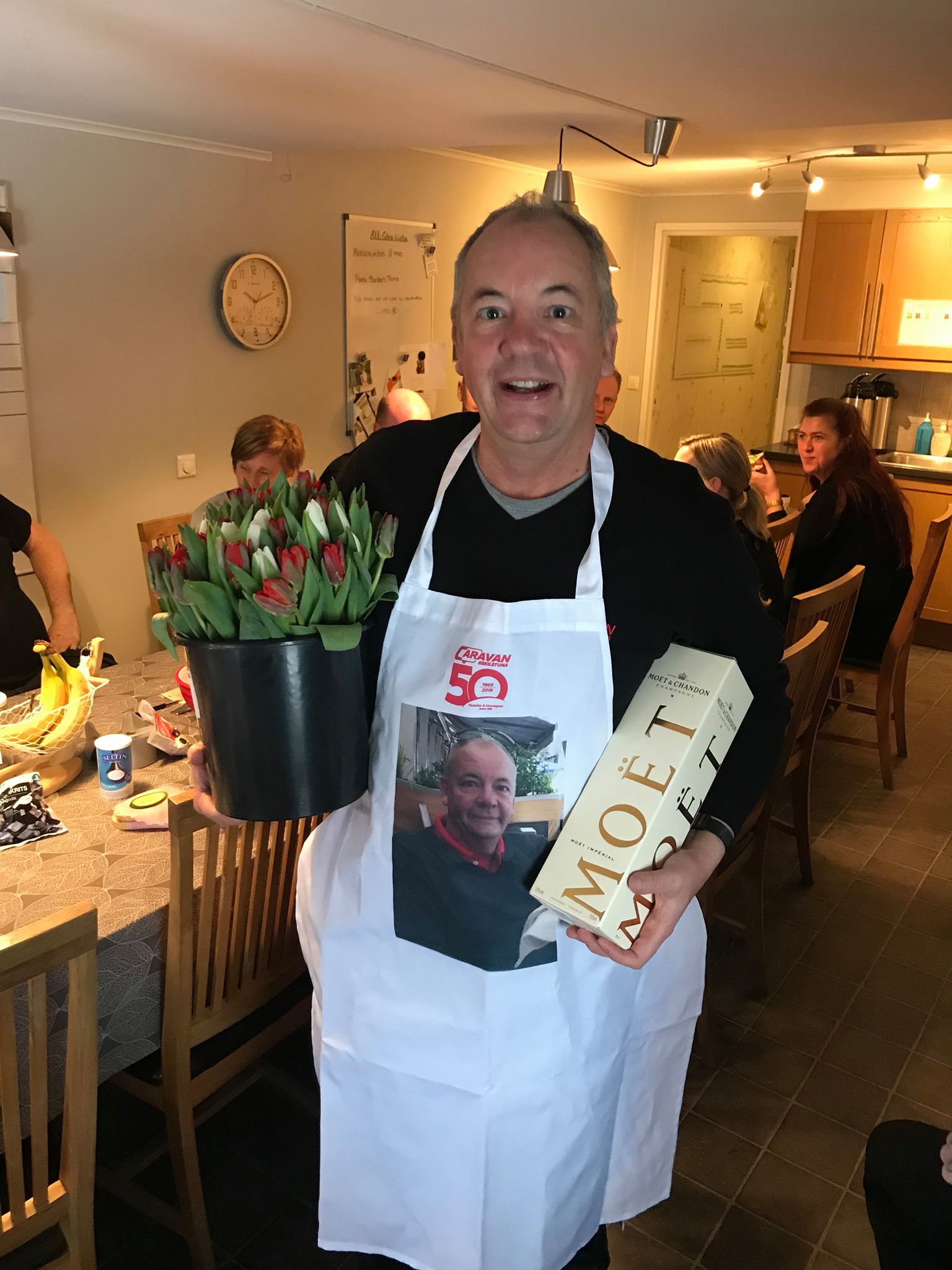 Man wearing apron with photo, holding flowers and champagne, smiling. Surrounded by people, in a kitchen.
