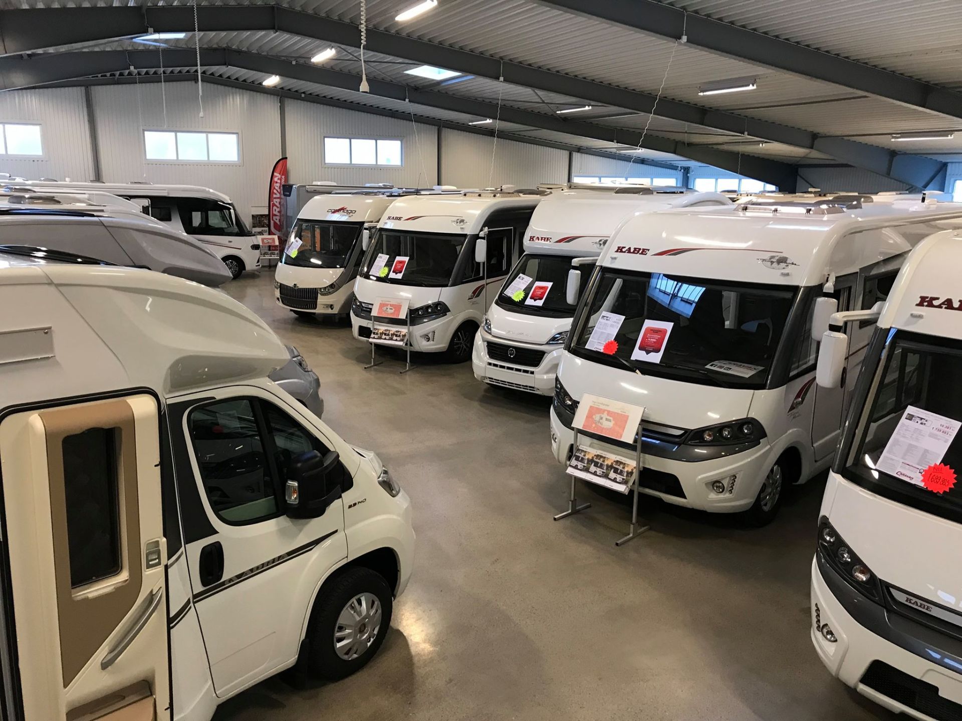 White RVs are parked inside a large warehouse. Some have price tags displayed.