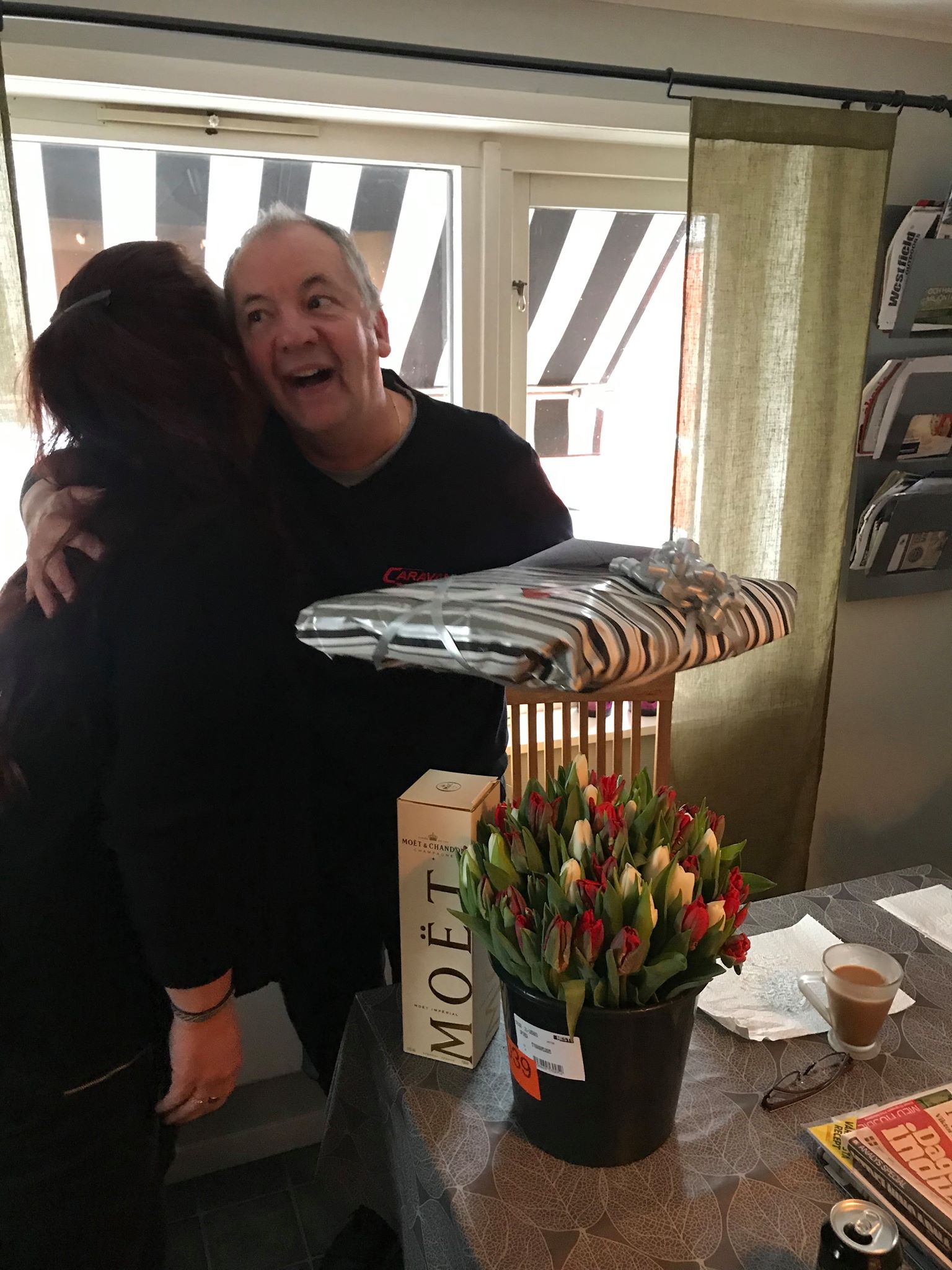 Man hugging person, smiling; model spaceship, flowers, and champagne bottle on table.