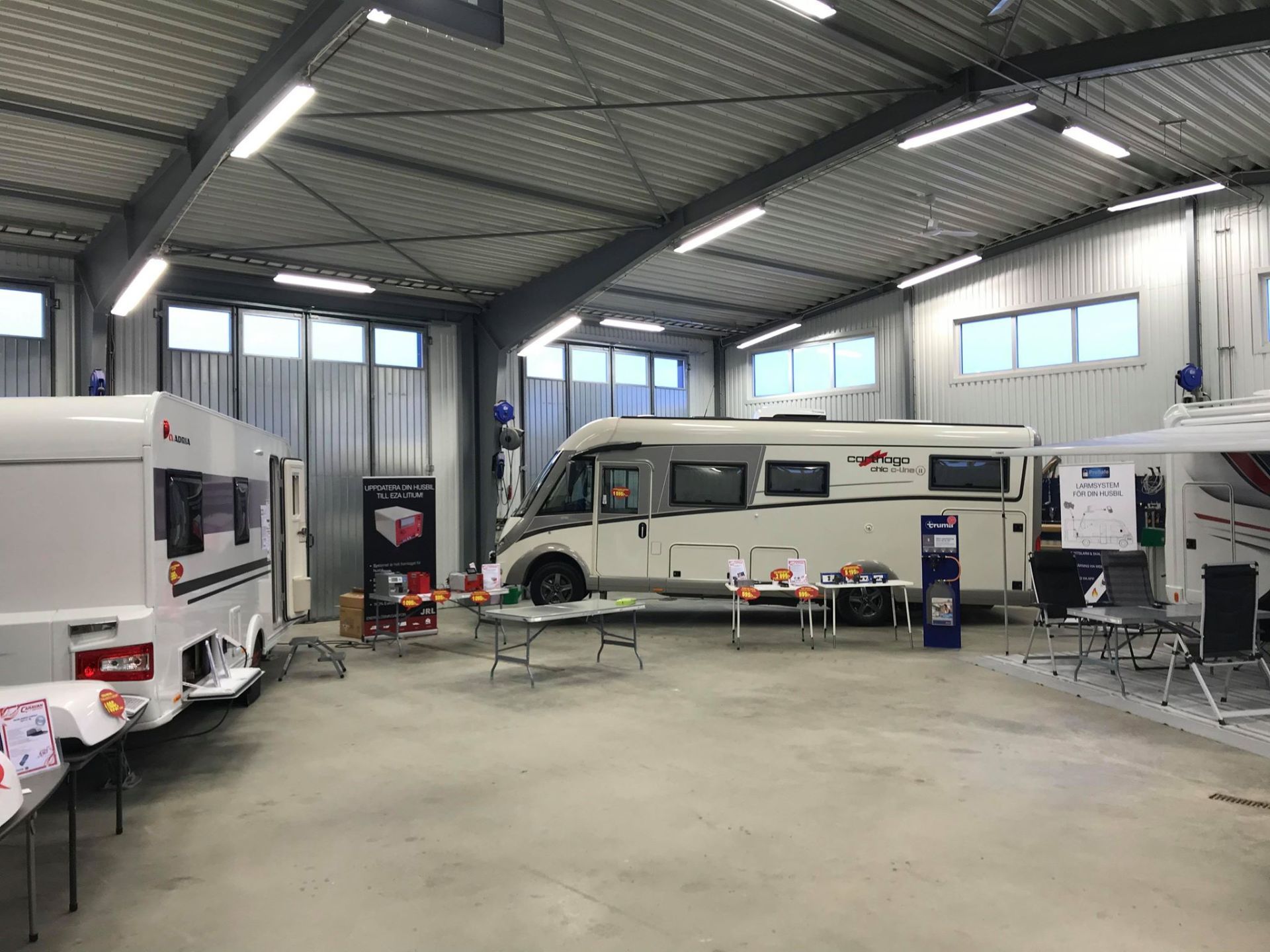 Large indoor space displaying recreational vehicles; RVs, trailer, tables, and product displays.