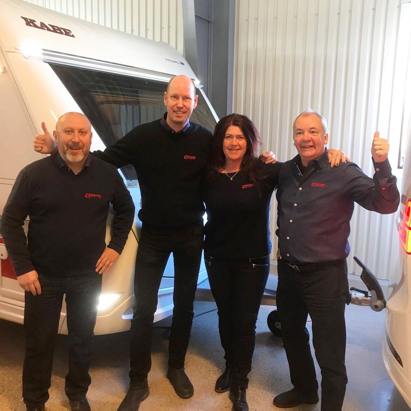 Four people smiling, giving thumbs up, pose by a white travel trailer in a garage.