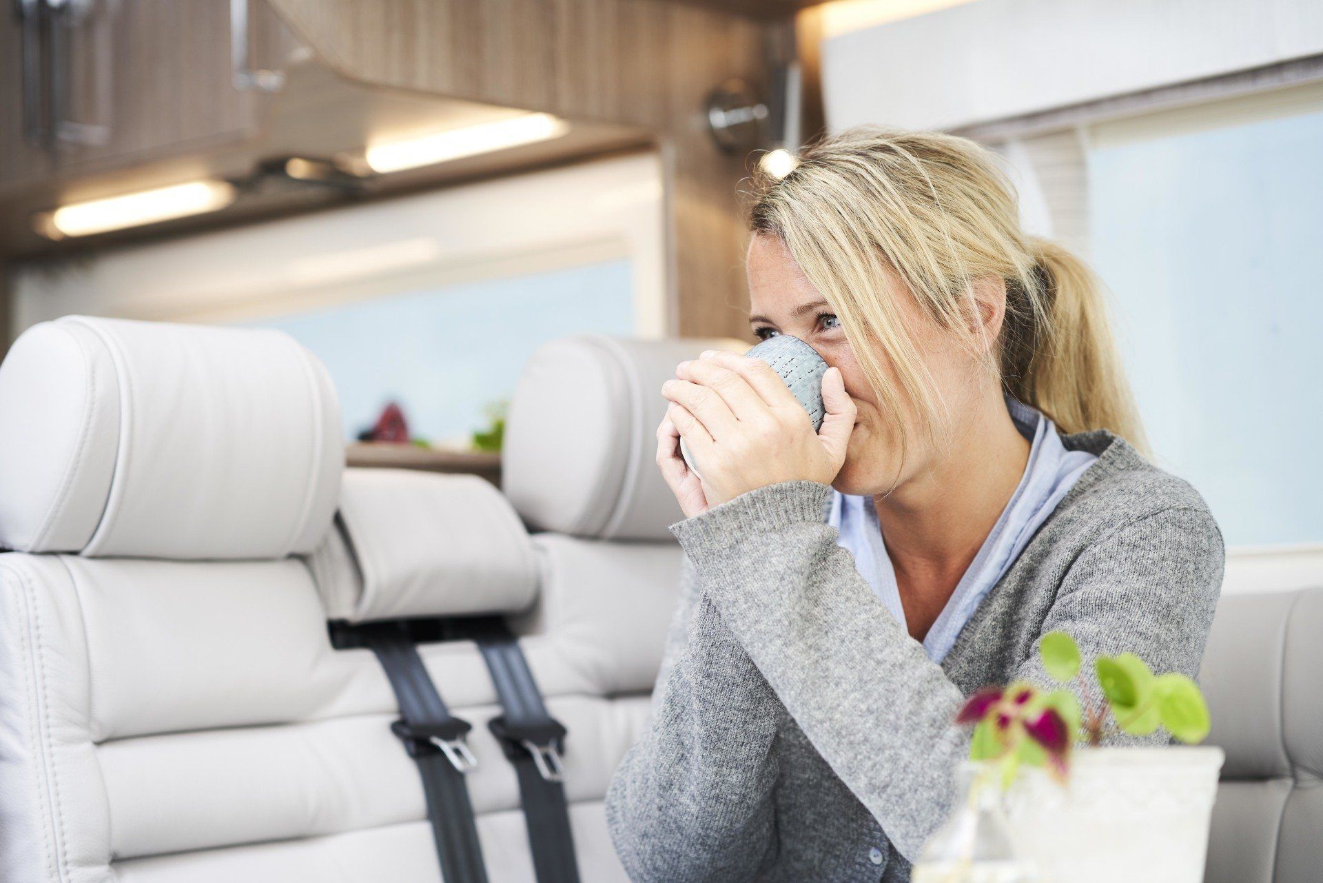 Woman inside a camper drinking from a mug, sitting near a white seat with black seatbelts.