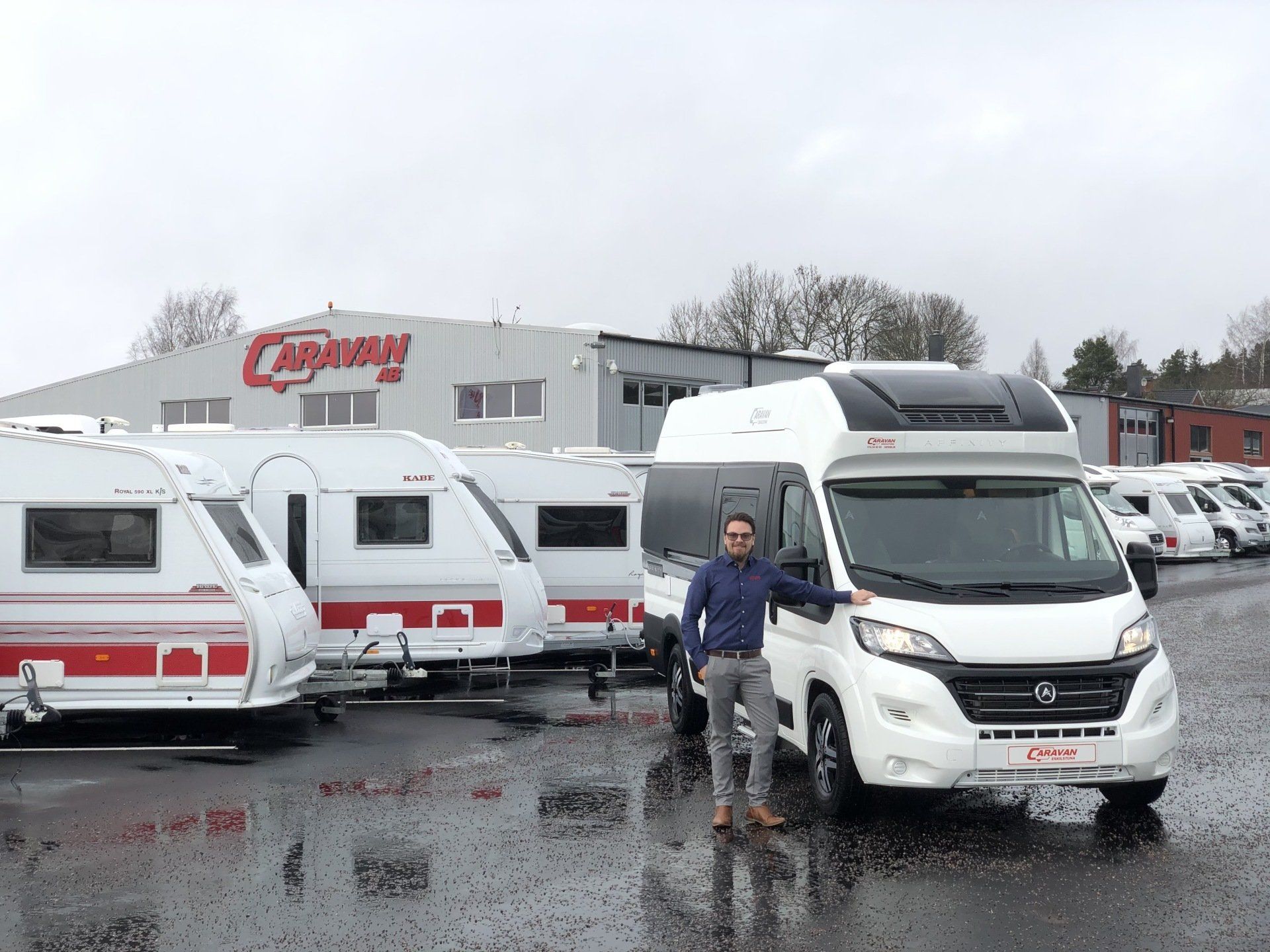 Man standing by white campervan in front of Camp-o-rama building. Several RVs are present.