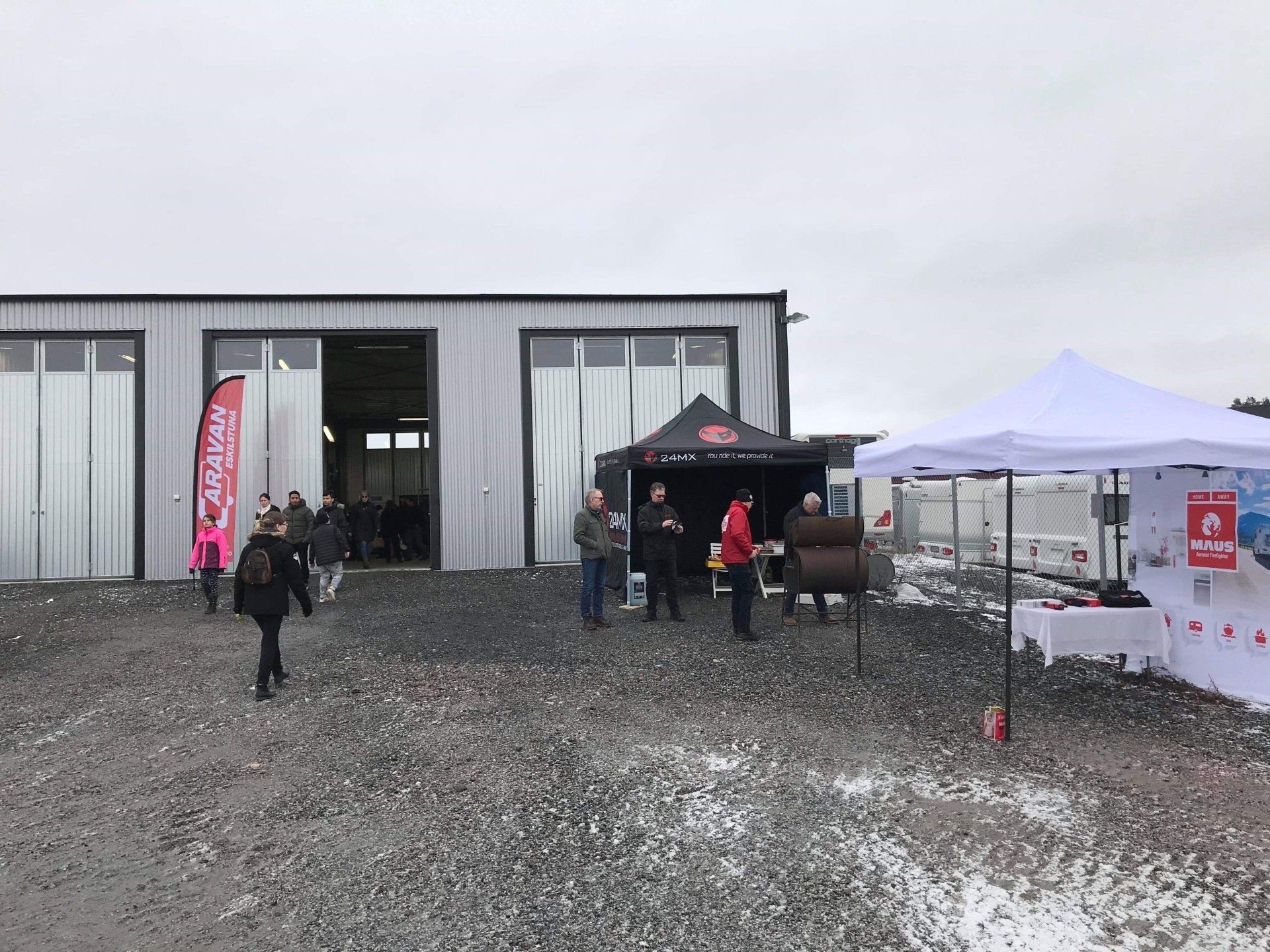People gathered outside a building with open garage doors and tents; gravel ground.