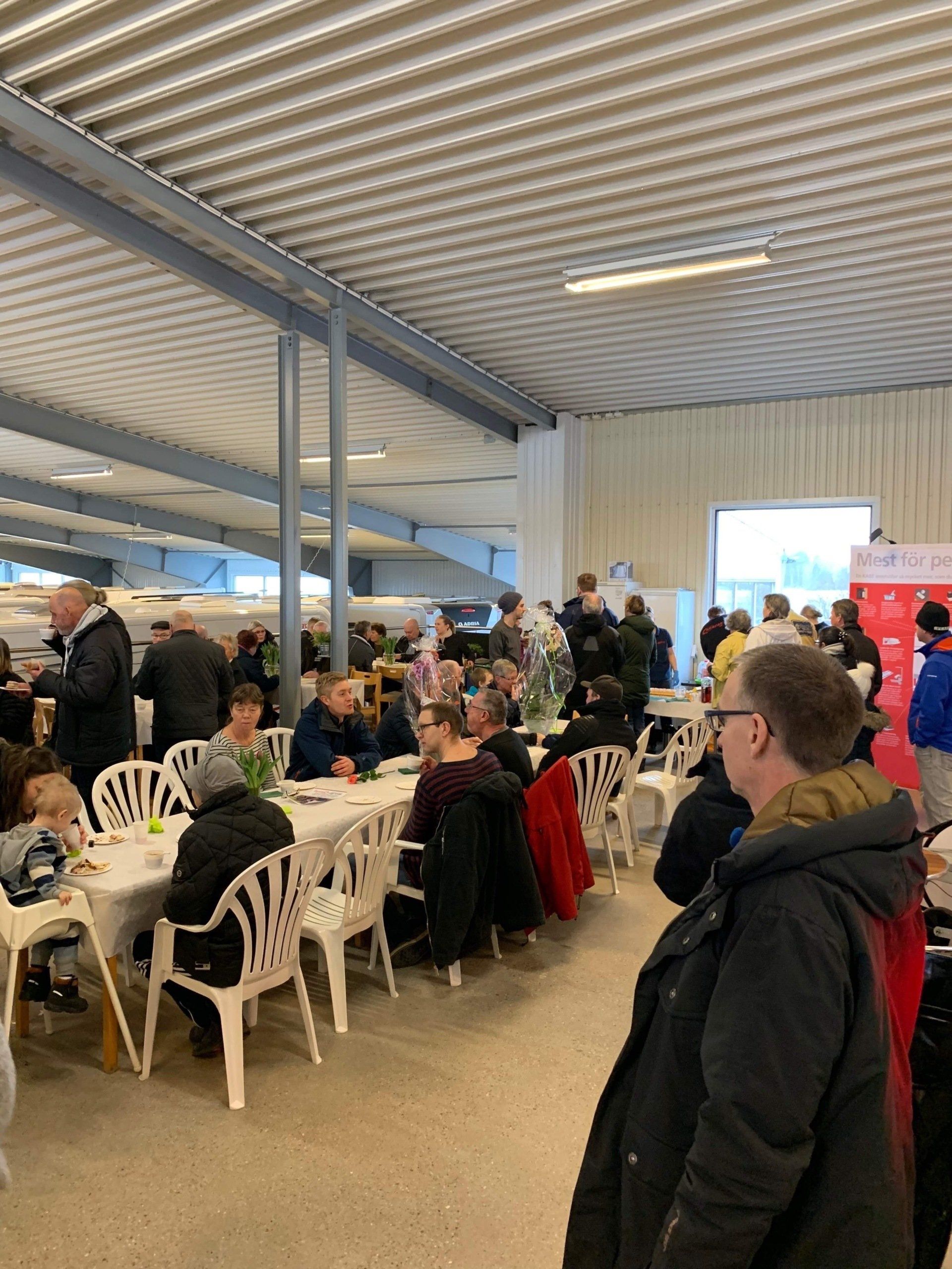 People seated at tables in a large, bright hall; some eating, others talking.
