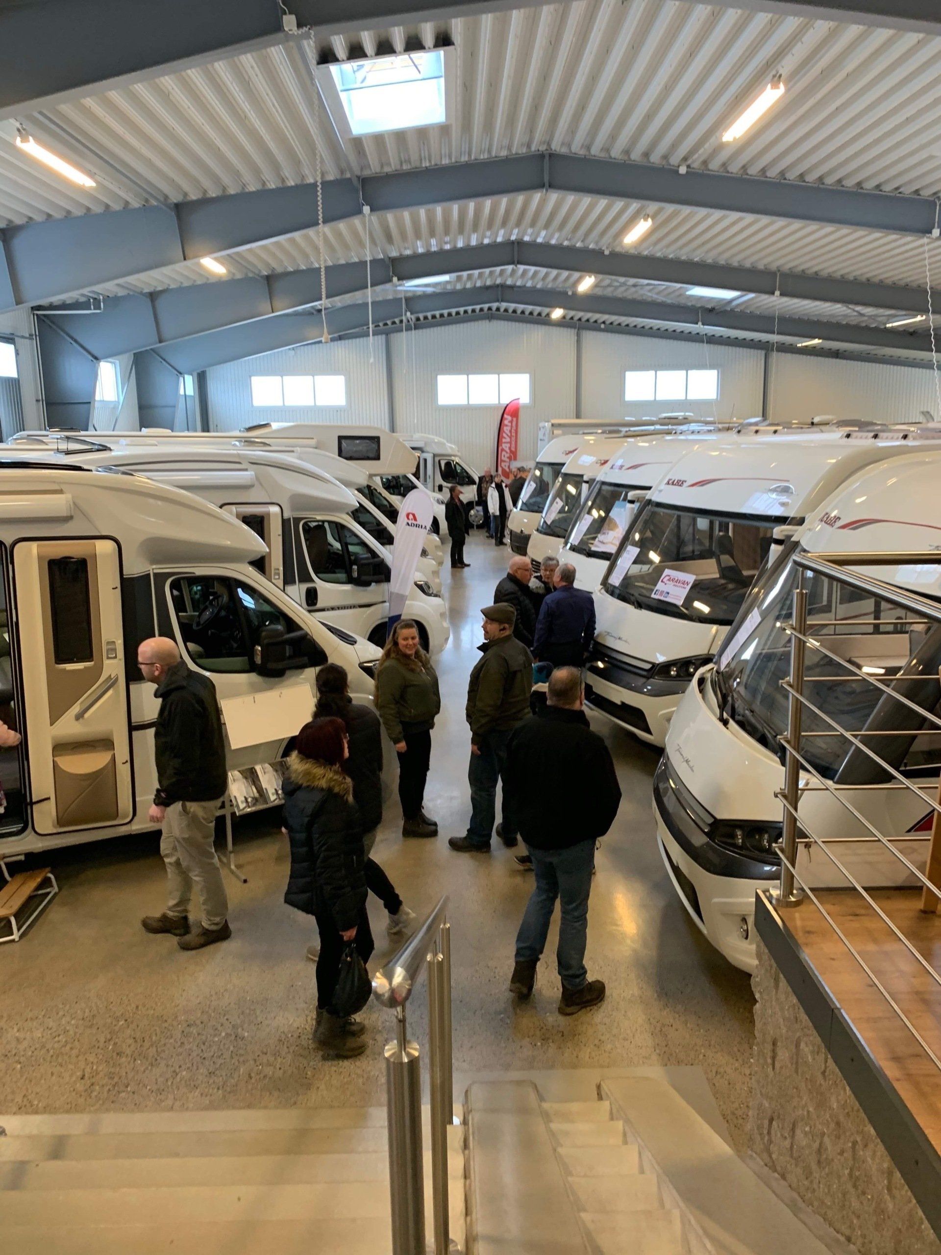 Inside RV showroom; people browsing white motorhomes under bright lights.