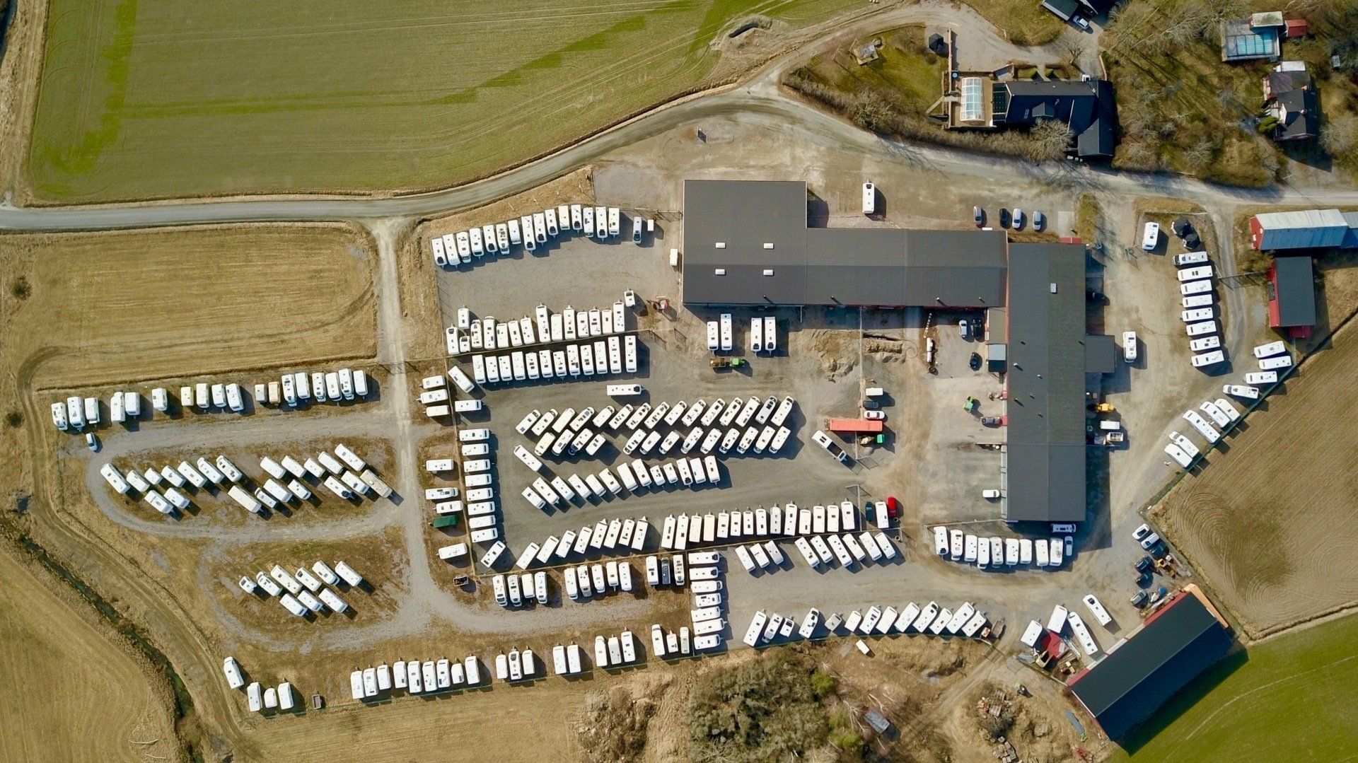 Aerial view of a vehicle storage lot with rows of white campers and a large commercial building.