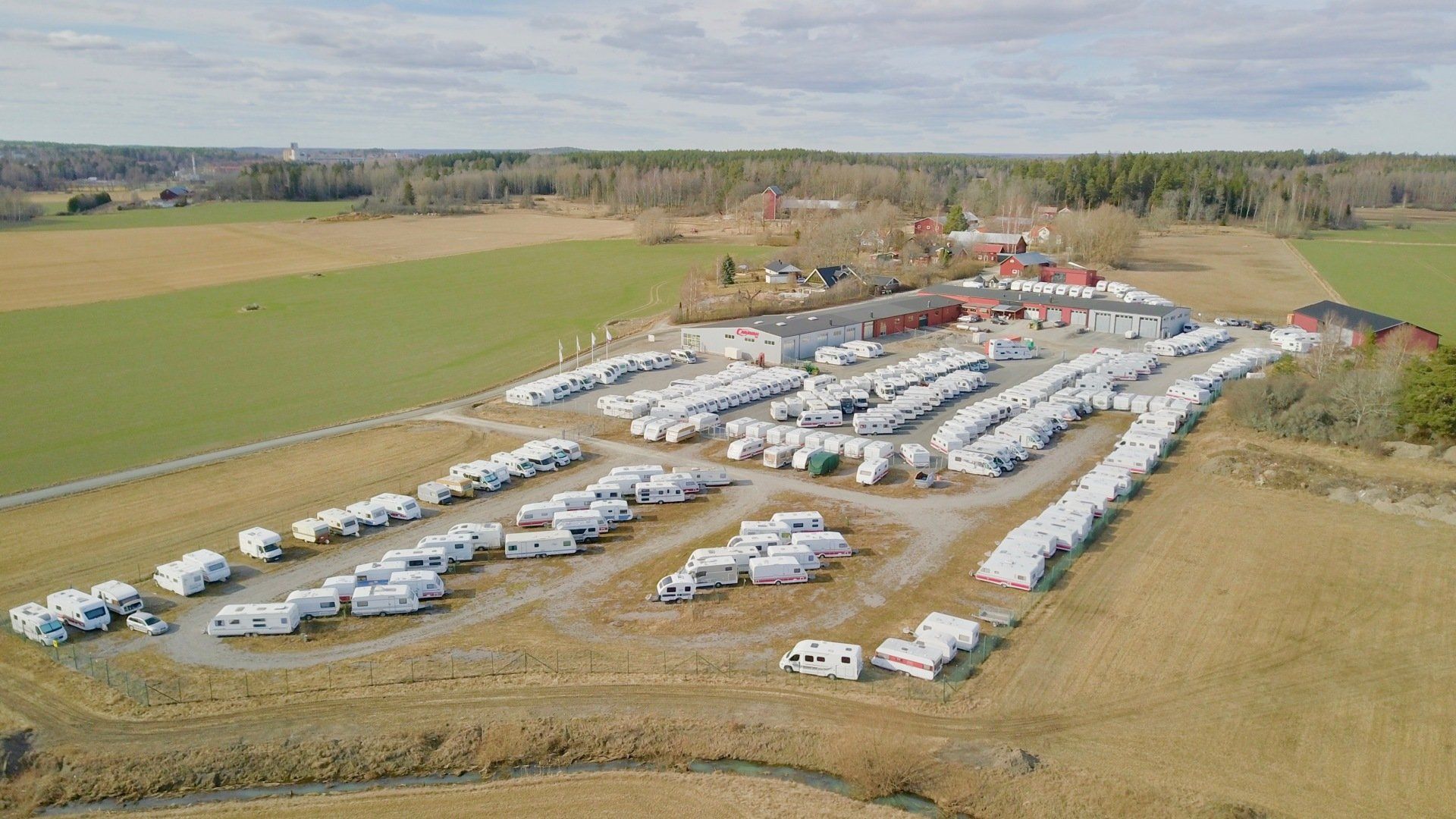 Aerial view of a lot filled with numerous white recreational vehicles parked on gravel, surrounded by fields and trees.