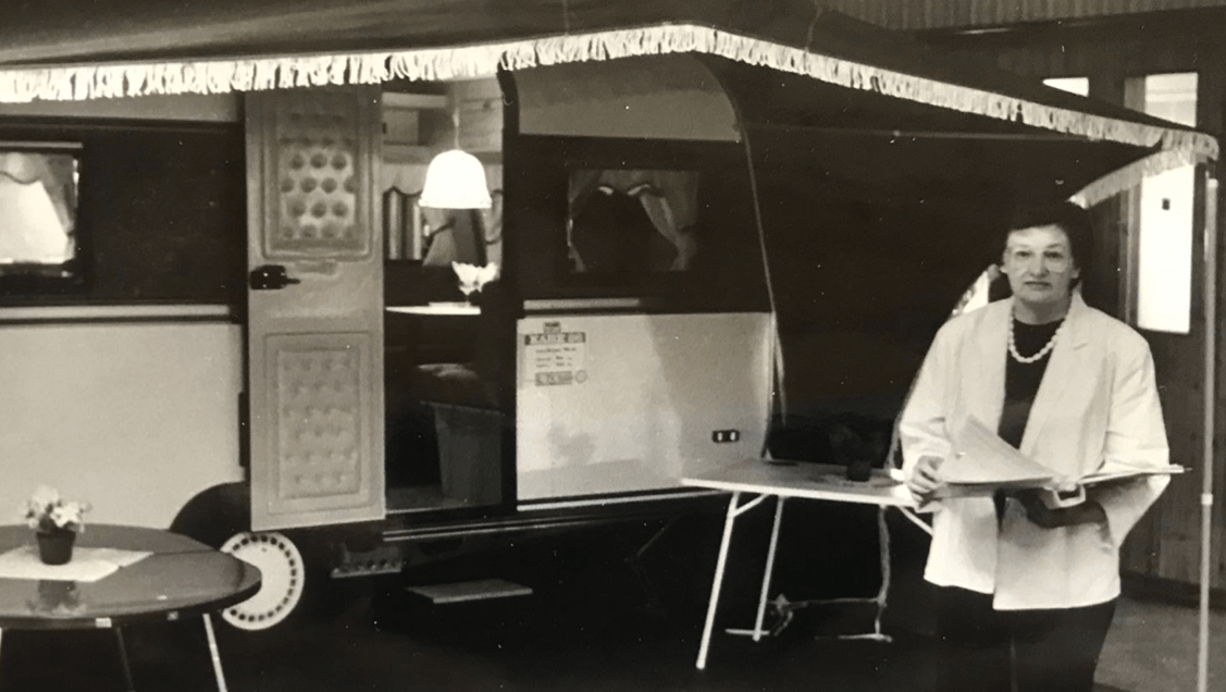 Woman stands next to a vintage caravan. It has a fringed awning, table. The woman holds papers.