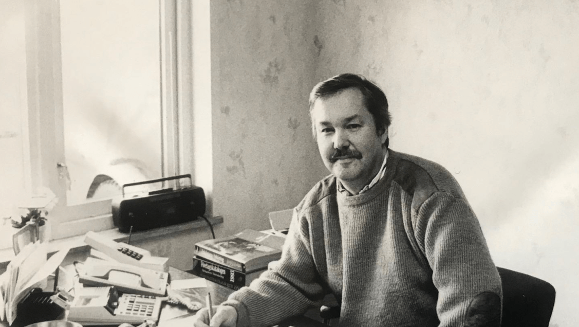 Man with a mustache sits at a cluttered desk near a window. He wears a sweater and looks toward the camera.