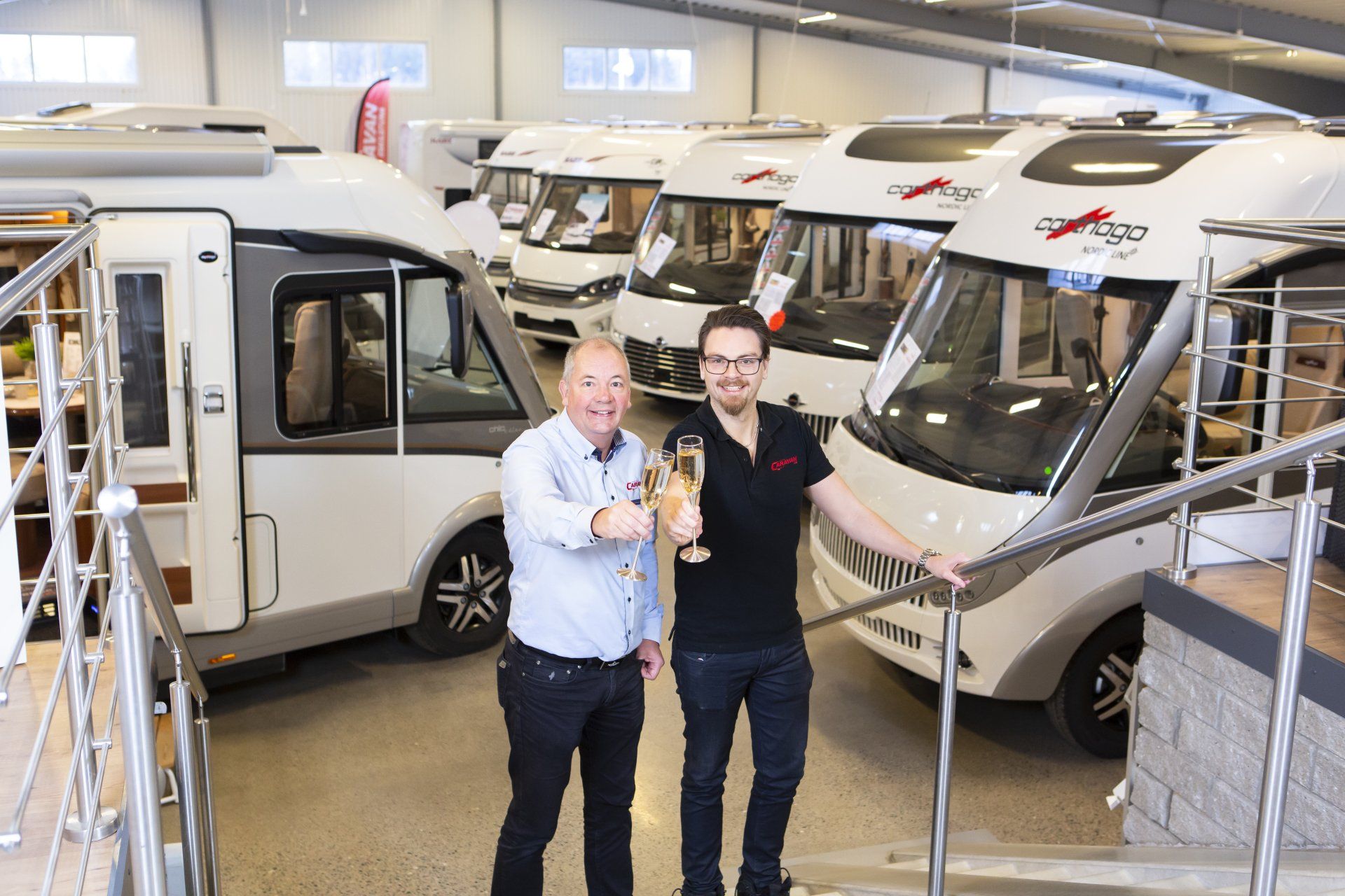 Two men toasting champagne in a showroom with several RVs.