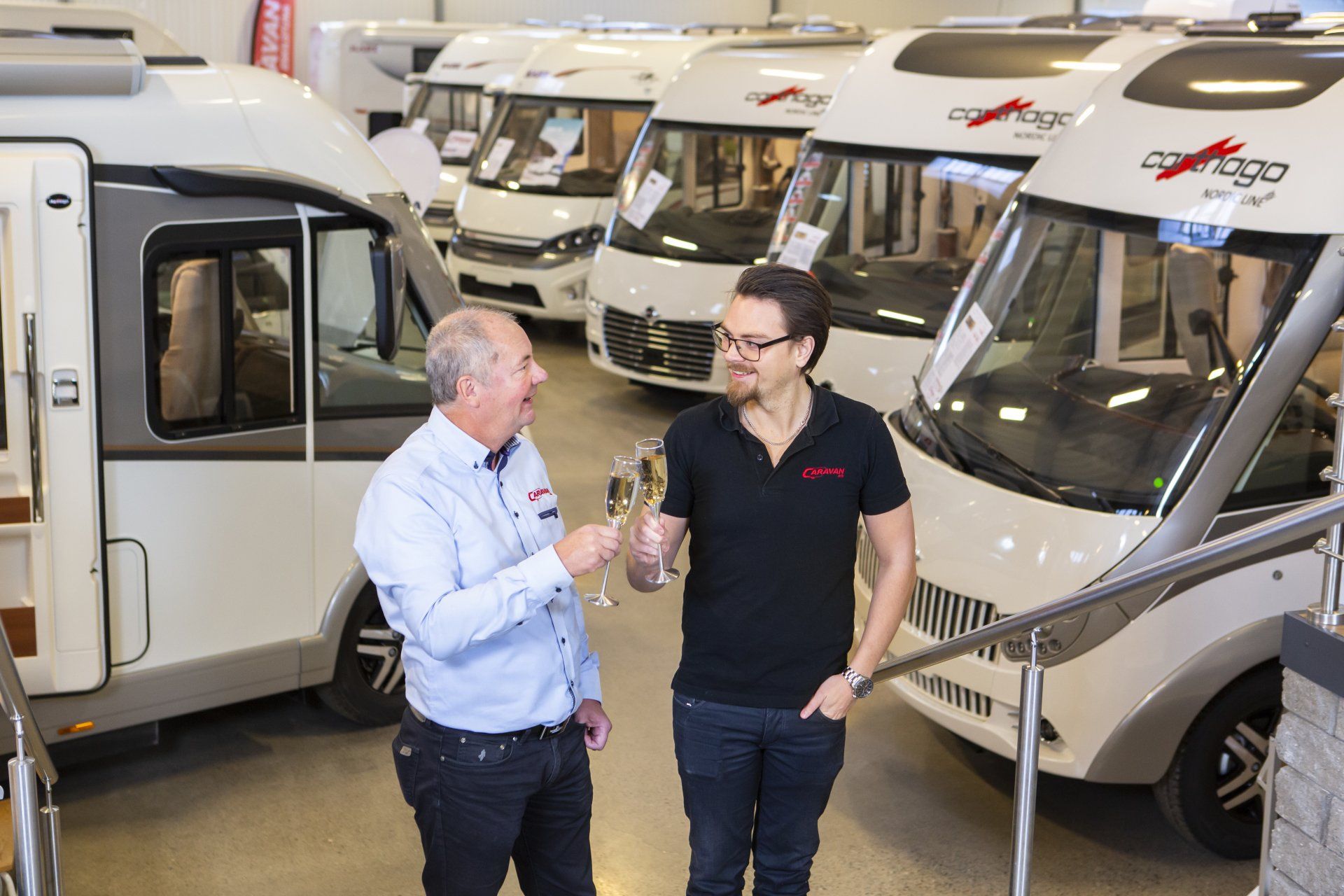 Two men toasting in a showroom with several camper vans. One holds a glass, smiling.