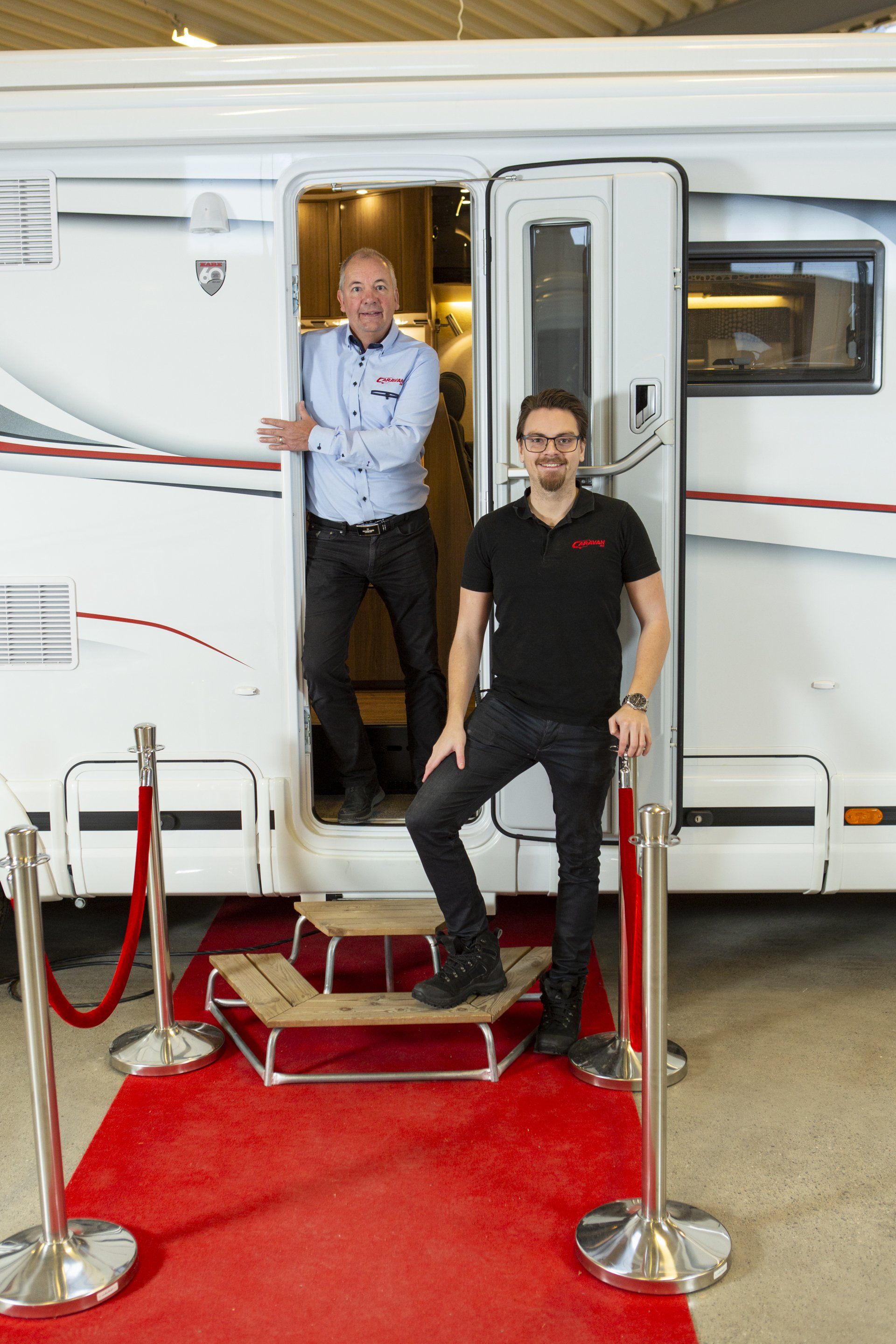 Two men stand by the open door of a motorhome on a red carpet.