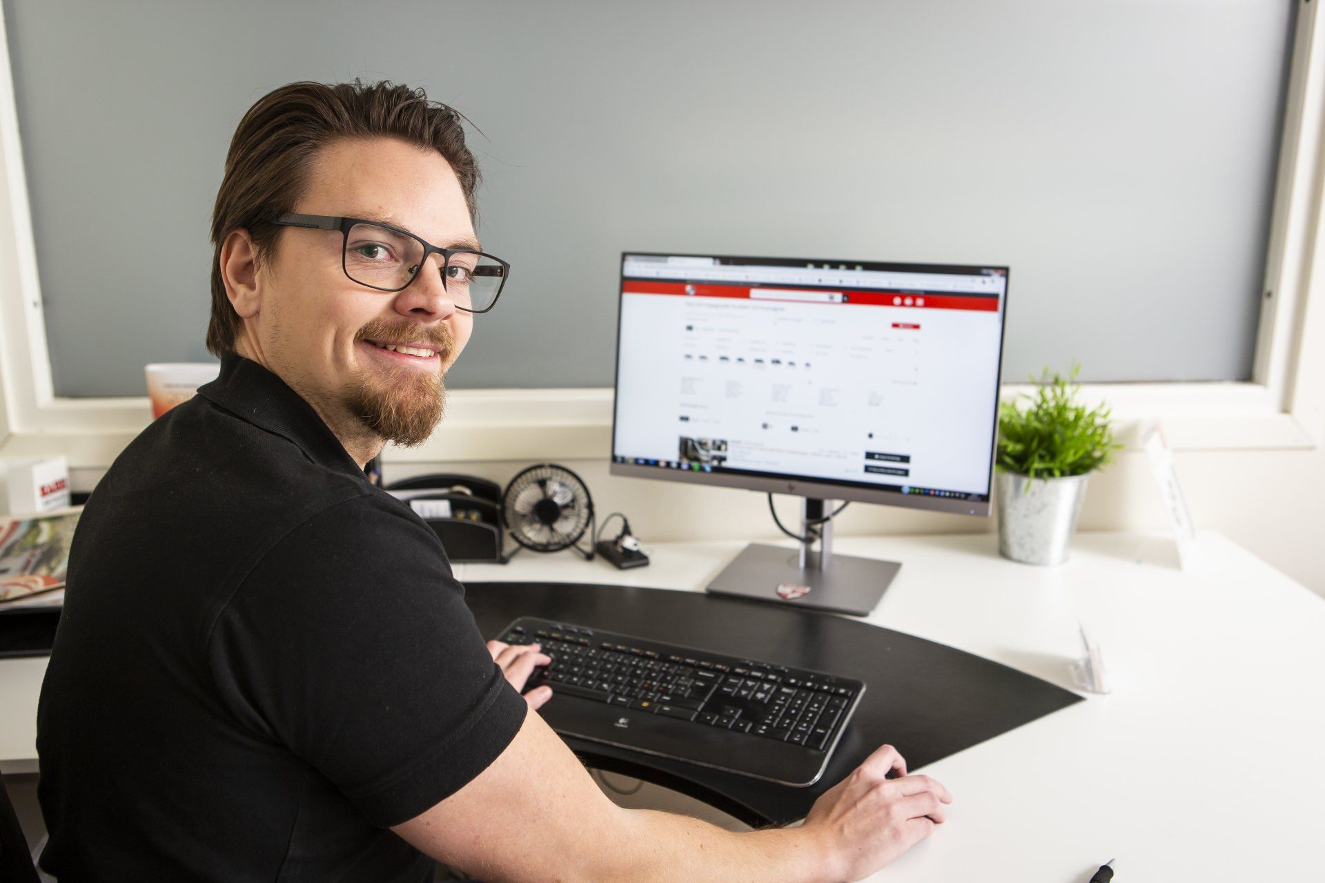 Man with glasses at a desk, looking at the camera and smiling. A computer monitor and keyboard are visible.