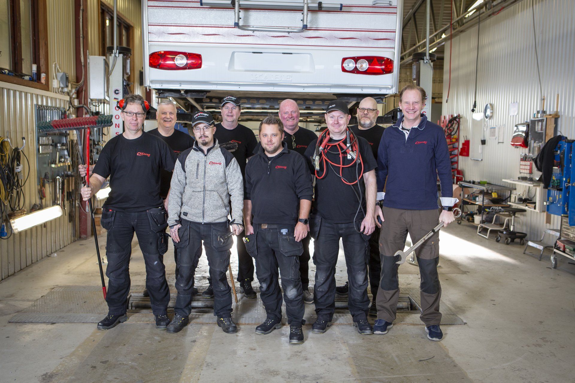 Group of mechanics in a garage, standing under a lifted RV. Several wear work clothes, smiling.