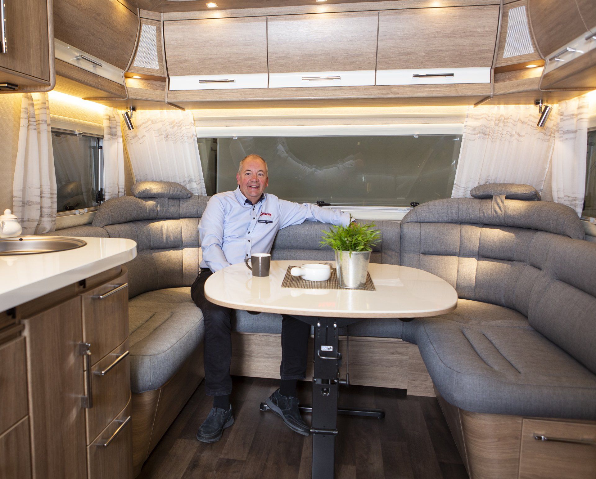 Man seated in a camper interior, smiling, near a table with plant and mug. Gray seating, wooden cabinets.