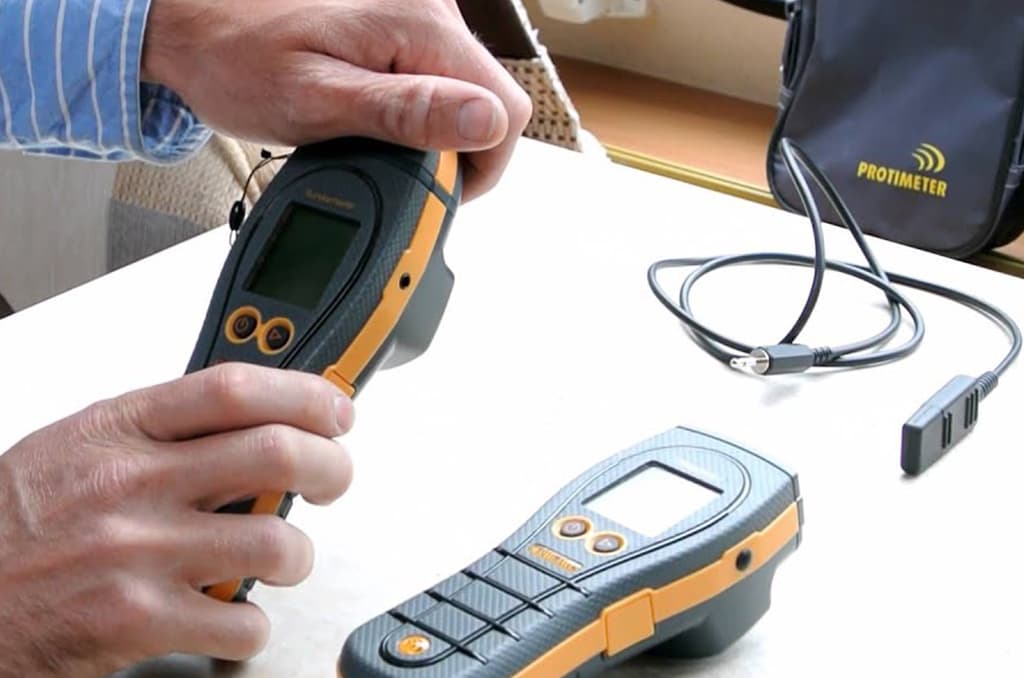 Person holding a yellow and gray moisture meter next to another meter, cord, and carrying case.